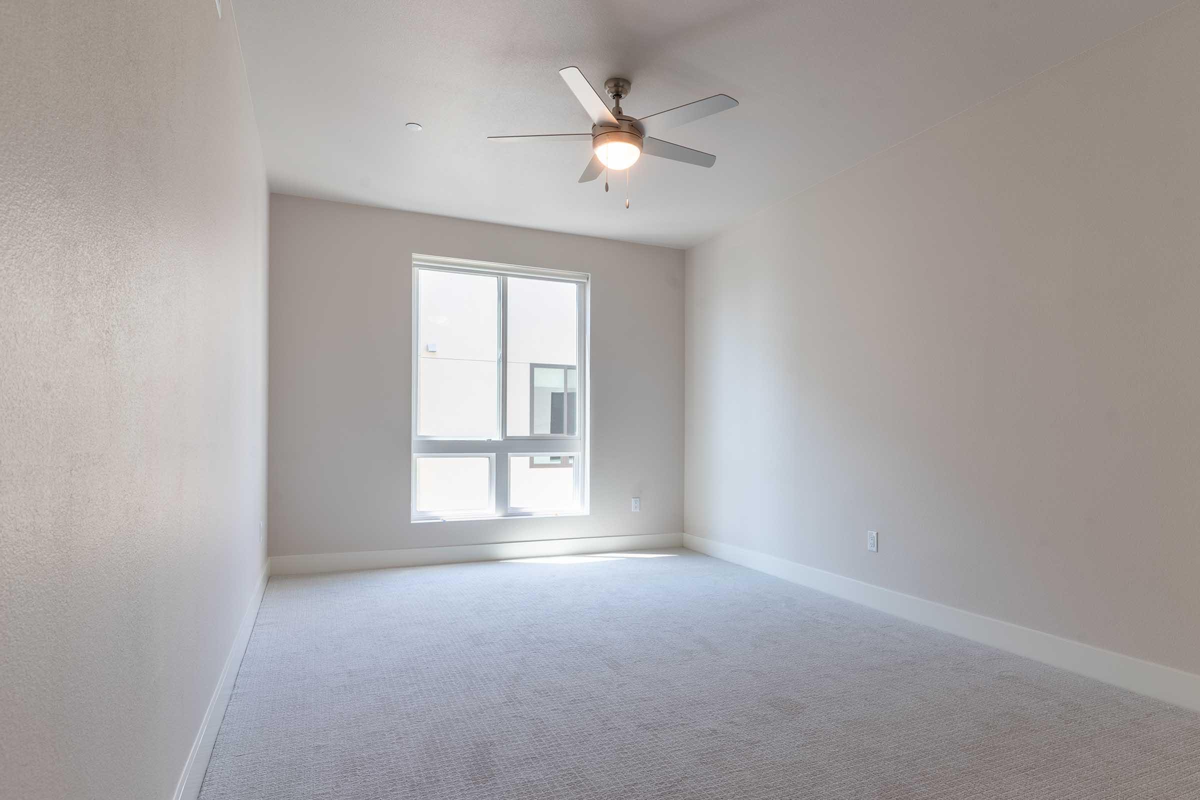 A bright, empty room with light gray walls and carpet. It features a ceiling fan and a large window allowing natural light to enter. There are no furnishings or decorations, creating a spacious and minimalist atmosphere.