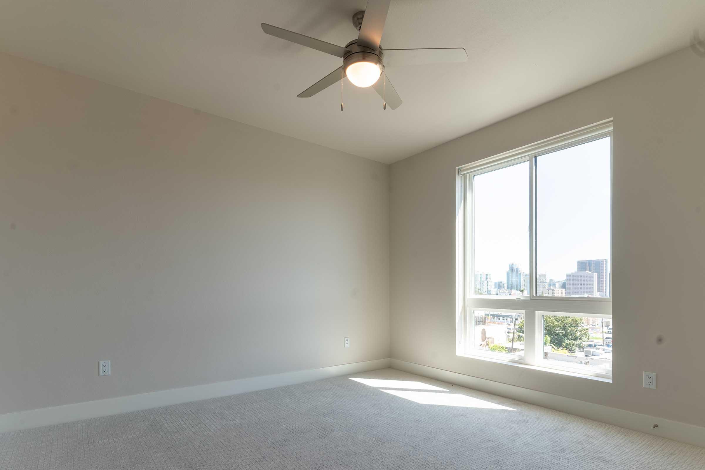 Empty room with light beige walls and carpet. A ceiling fan with a light fixture is mounted in the center. A large window lets in natural light, showcasing a cityscape outside. The space appears bright and fresh, ideal for decoration or furnishing.