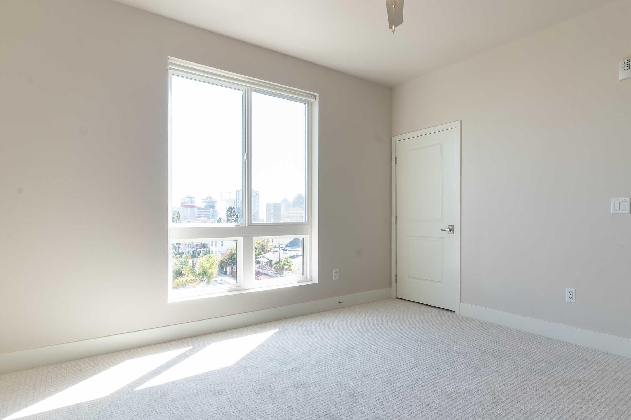 Bright, empty room with light-colored walls and a carpeted floor. A large window lets in natural light, offering a view of buildings outside. The door is closed, and there are no furnishings in the space, creating a clean and open feel.