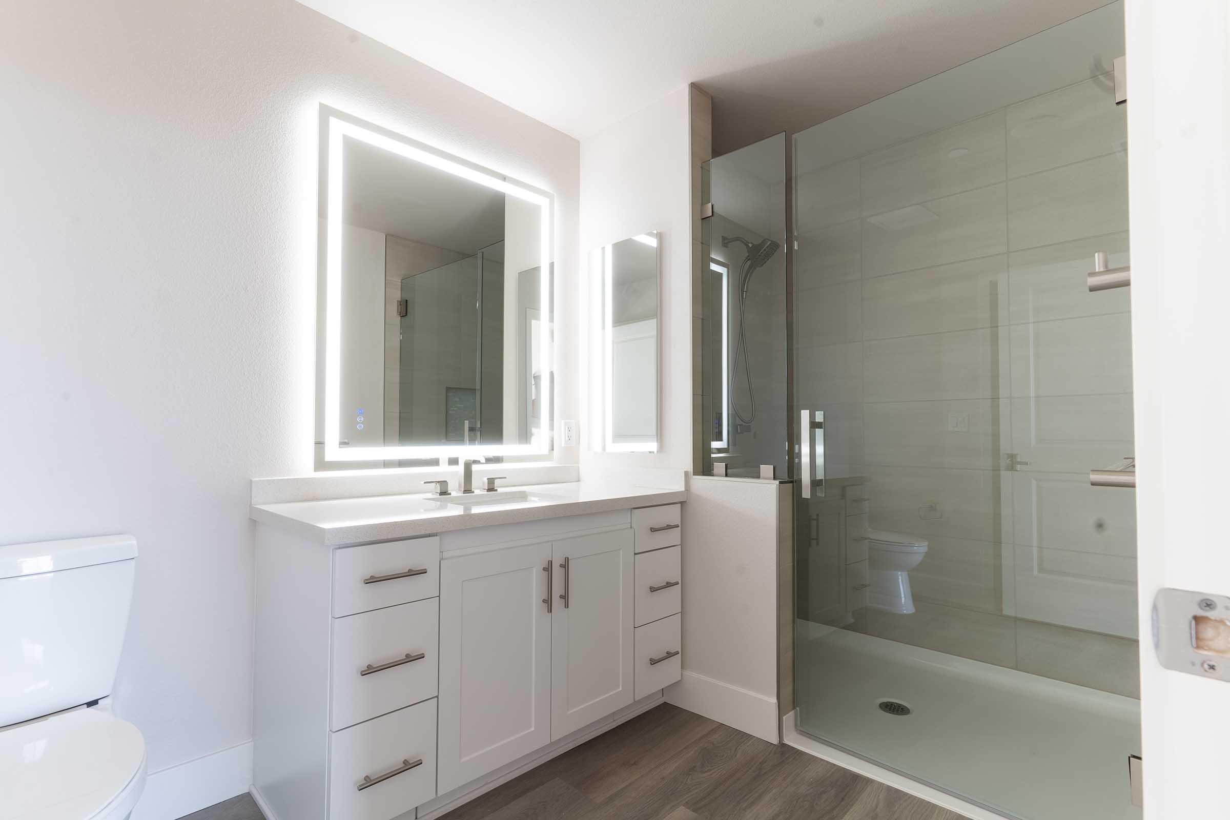 Modern bathroom featuring a large glass shower, vanity with dual sinks, and backlit mirrors. The design is clean and minimalist, with light-colored walls and flooring, providing a spacious and bright atmosphere. A toilet is positioned separately, enhancing the overall functionality of the space.