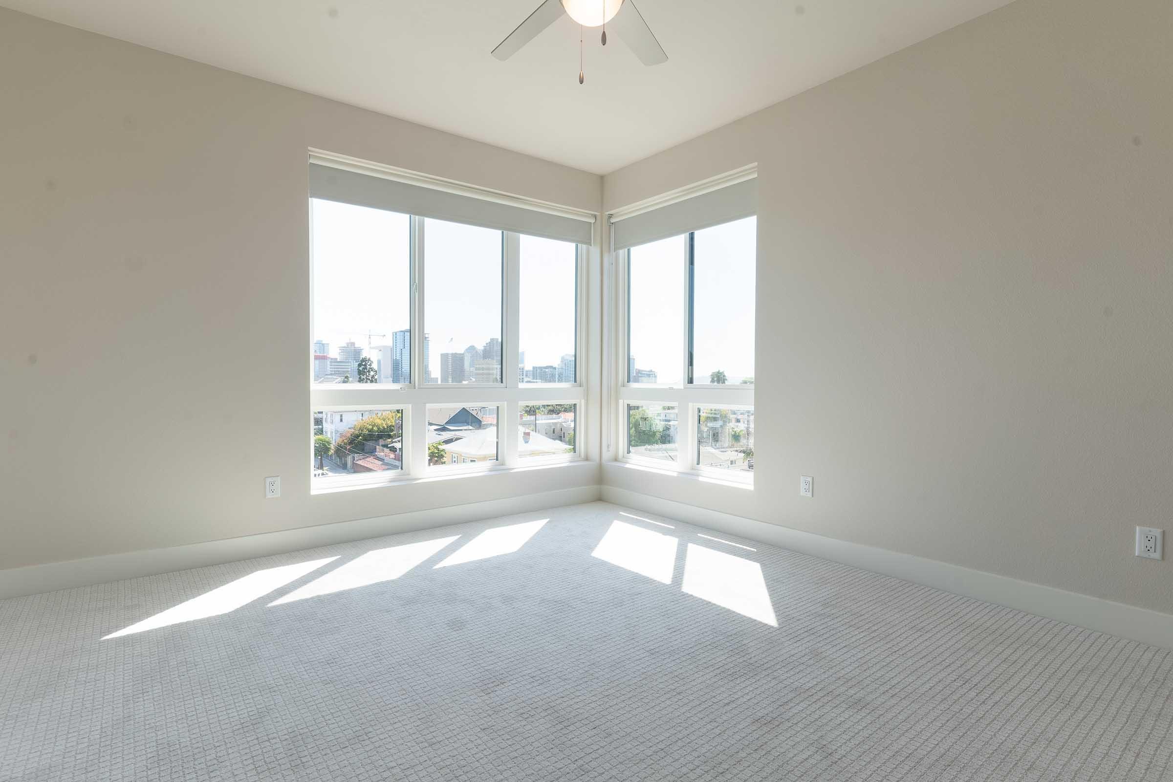 Empty room with light-colored walls and a carpeted floor. Features large windows allowing ample natural light, offering a view of the city skyline. A ceiling fan is visible, and the overall atmosphere is bright and airy.