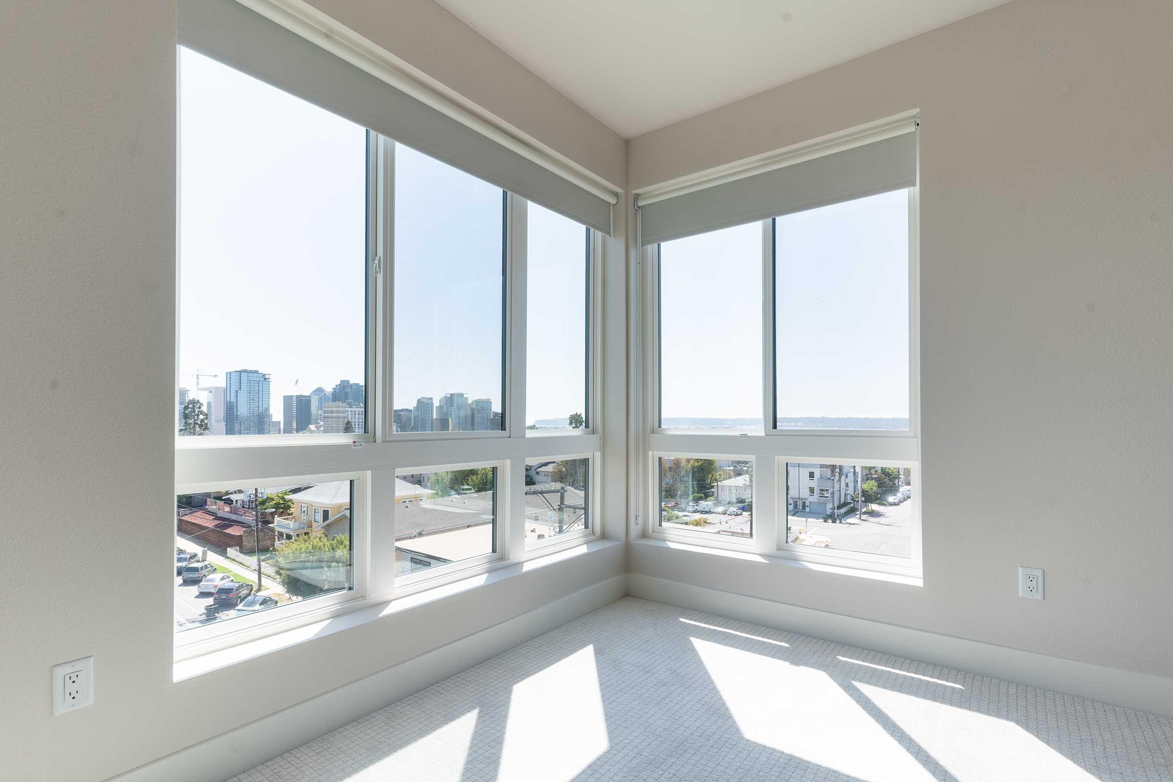 Bright, modern room with large windows offering a panoramic view of a city skyline. Natural light floods the space, highlighting the neutral-colored walls and carpeted floor. The room is cornered, providing a unique vantage point of the outdoor scene.