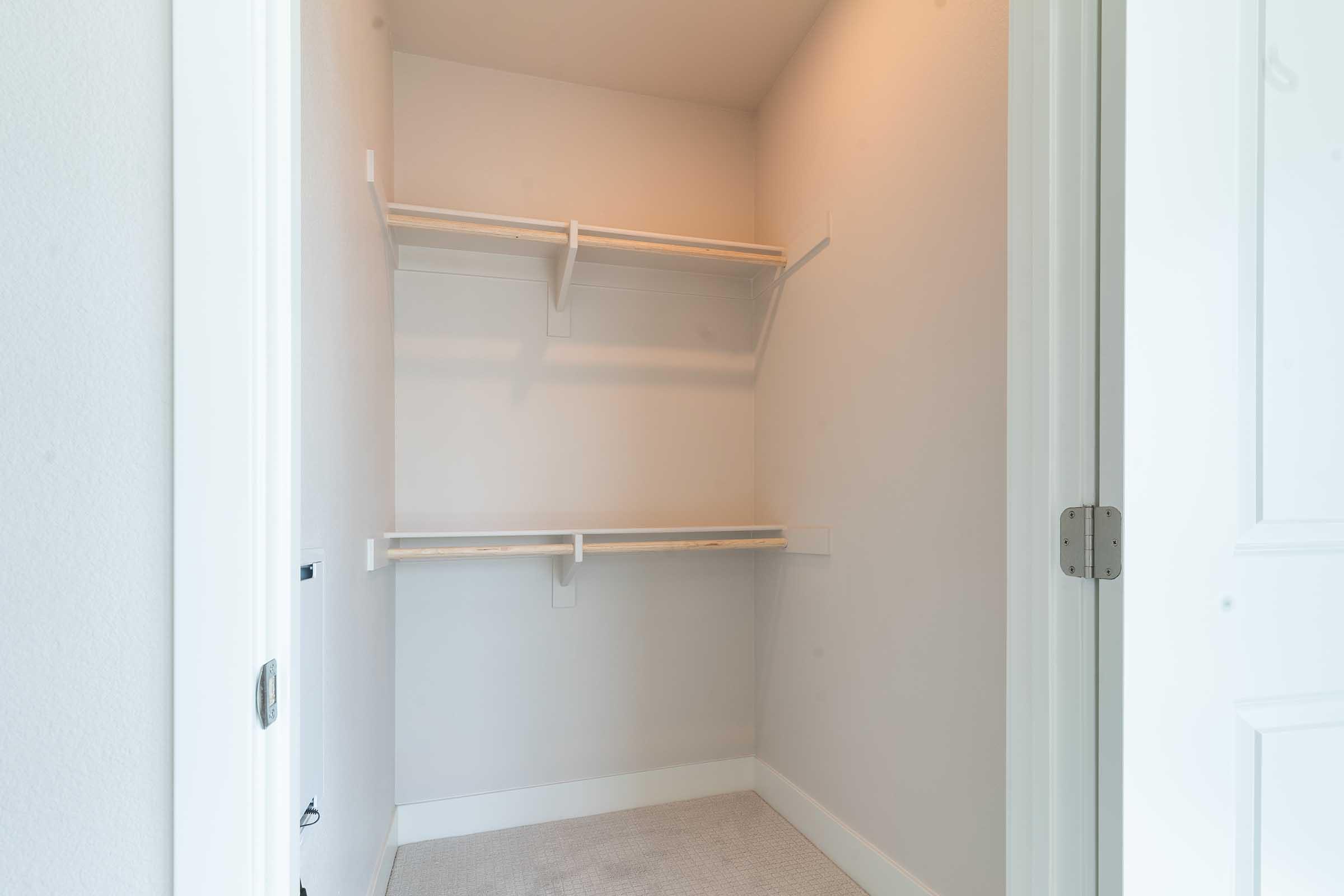 An empty closet with light-colored walls and two wooden shelves mounted on the wall. The floor is covered with a light carpet, and there is a white door partially open, showing simple door hardware. The space has a bright, airy feel due to the natural light.