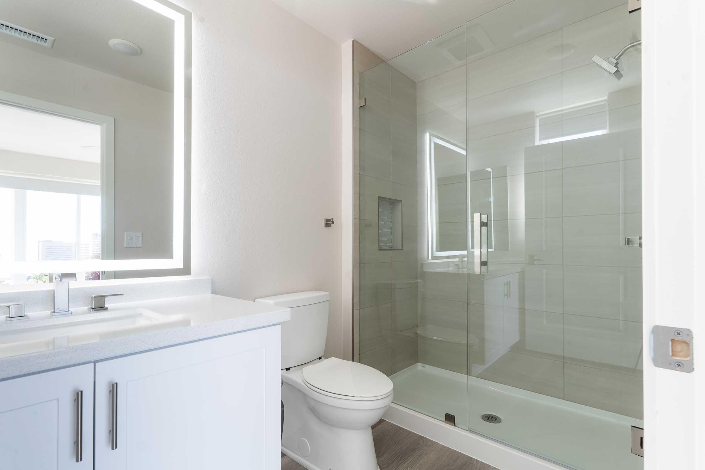 A modern bathroom featuring a glass shower enclosure, a white toilet, and a sleek vanity with two sinks. The space has neutral tile walls, ambient lighting from a backlit mirror, and large windows allowing natural light to enter.