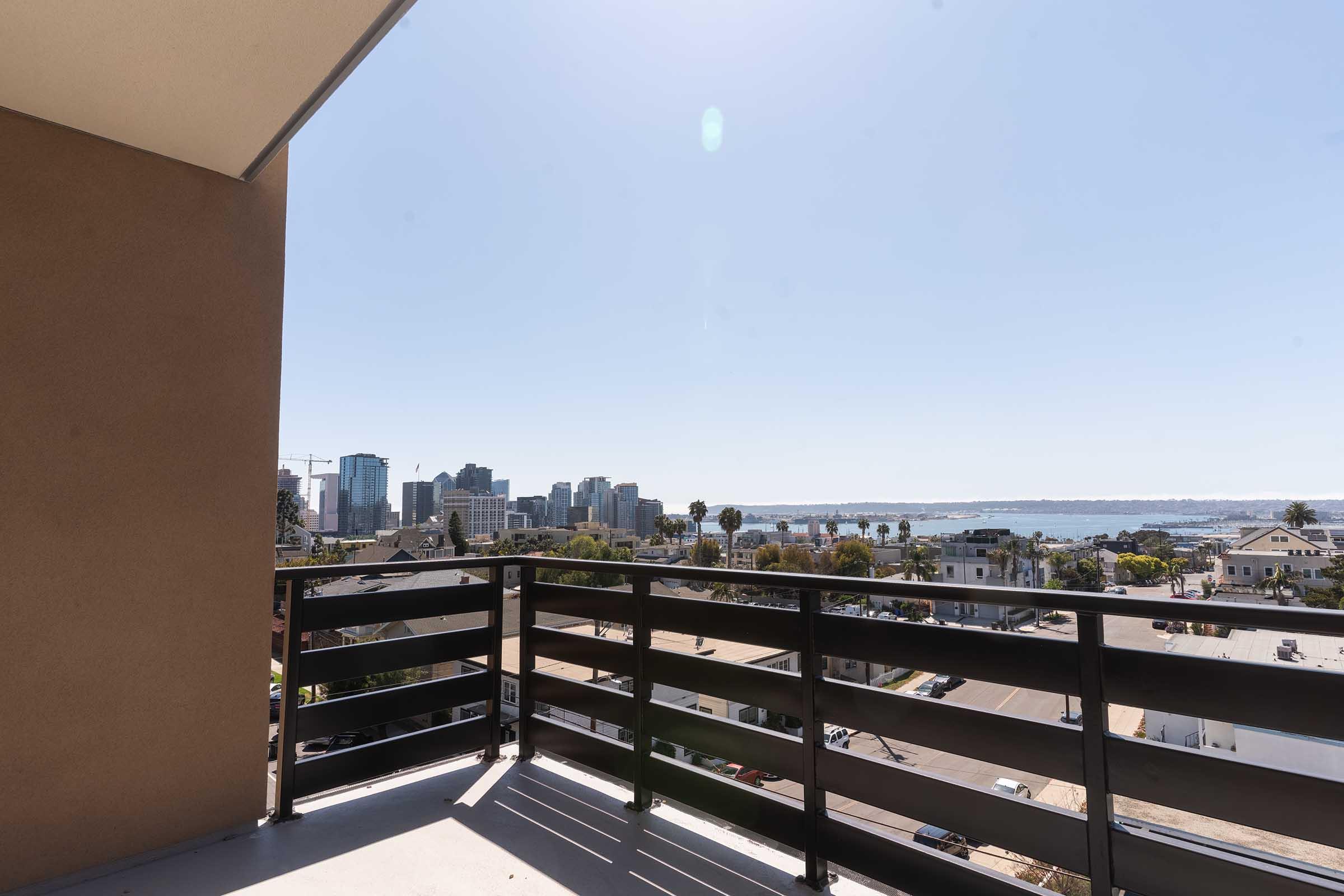 A bright, sunny balcony view overlooking a city skyline with modern buildings and palm trees, leading to a distant view of the water. Clear blue sky adds to the vibrant atmosphere, showcasing a mix of urban and natural elements.