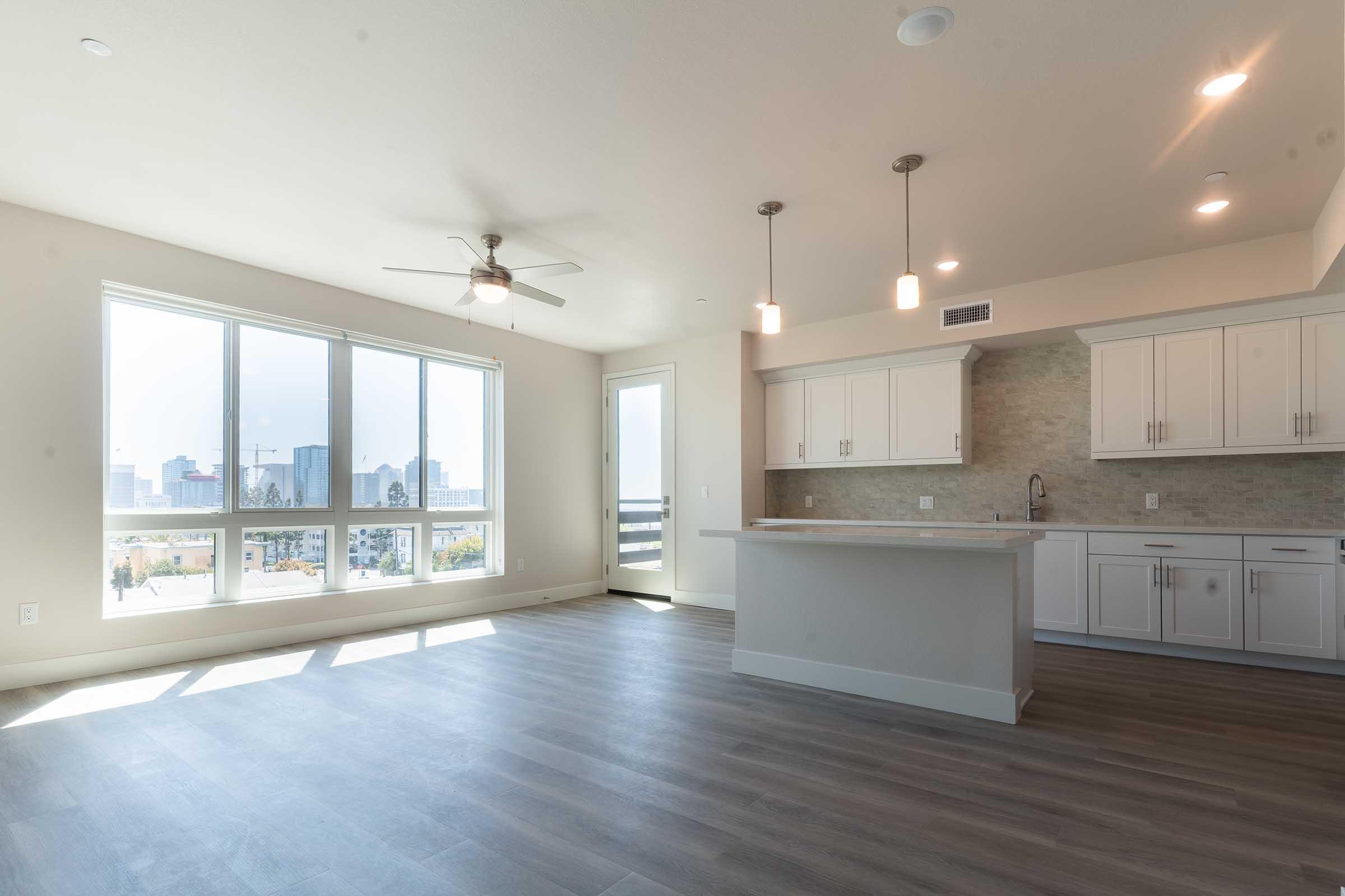 Spacious modern apartment interior with large windows allowing plenty of natural light. Features a kitchen with white cabinets, gray countertops, and a central island. Open floor plan leads to a living area with wooden flooring and a ceiling fan, offering a view of the city skyline.