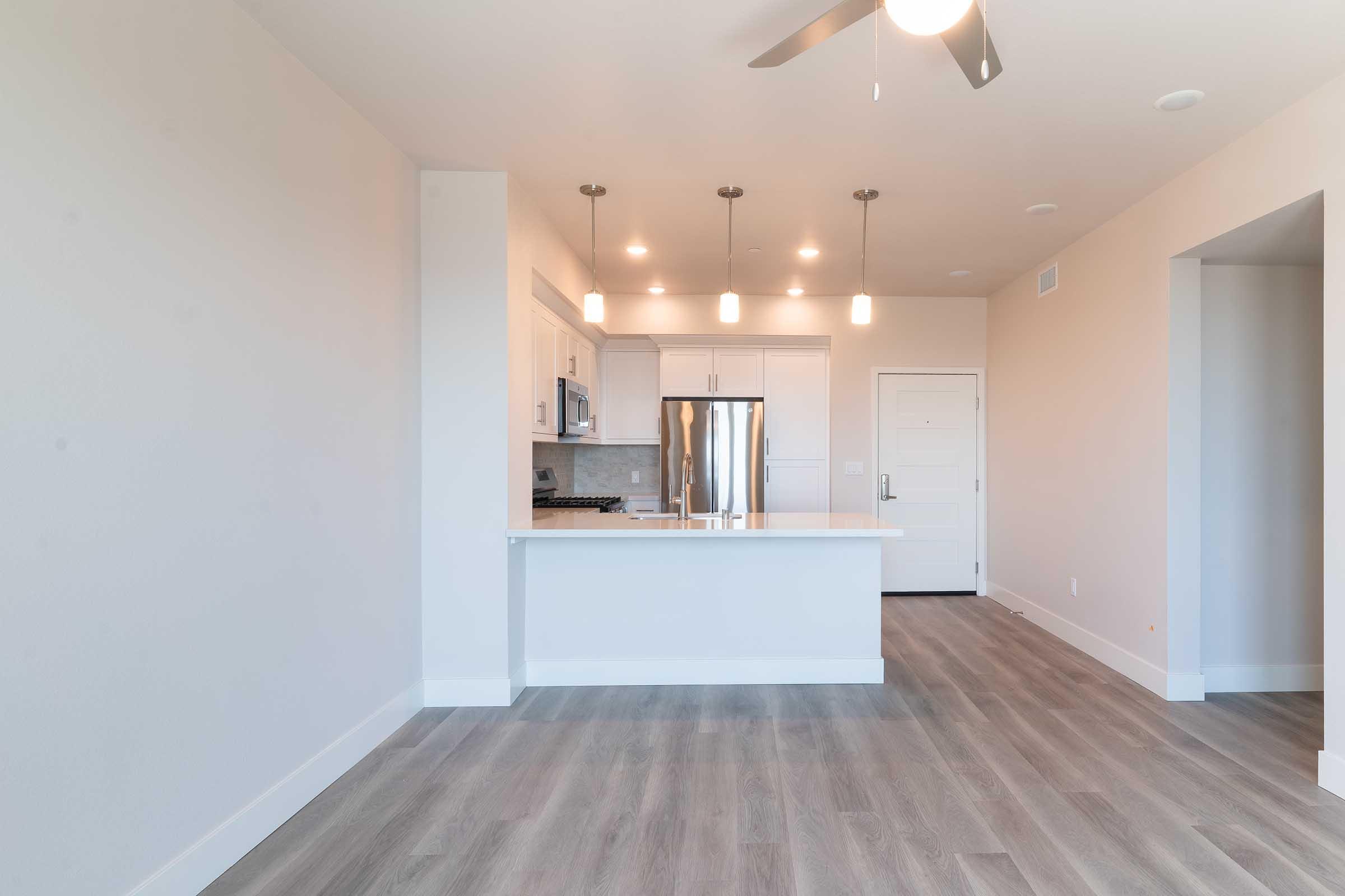 A modern kitchen and living area featuring a large island with a stainless steel refrigerator, pendant lighting, and sleek cabinetry. The space has light-colored walls and hardwood flooring, creating a bright and open atmosphere. The entrance door is visible on the right.
