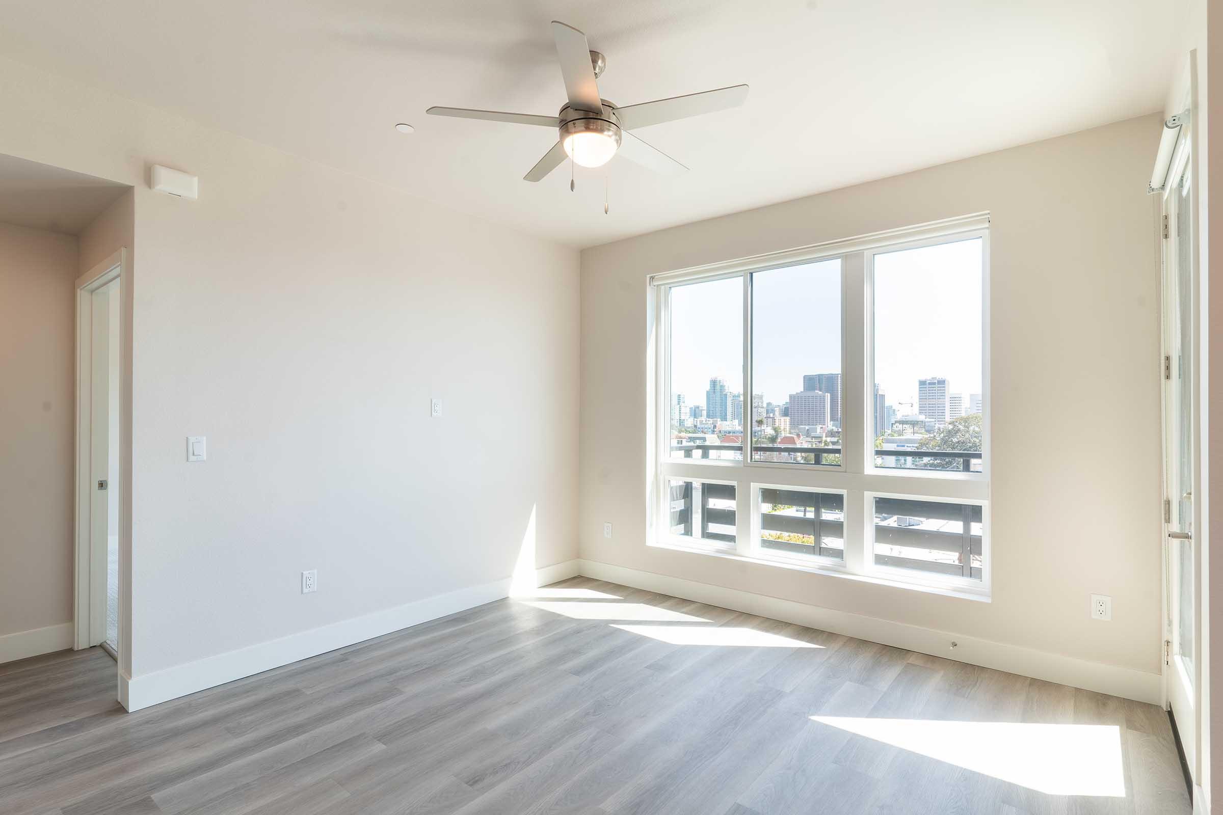 Bright, modern room with large windows showcasing a city view. A ceiling fan is installed, and the light-colored walls complement the light wooden flooring. The space is empty, offering a clean and airy atmosphere, perfect for personalizing and decorating.