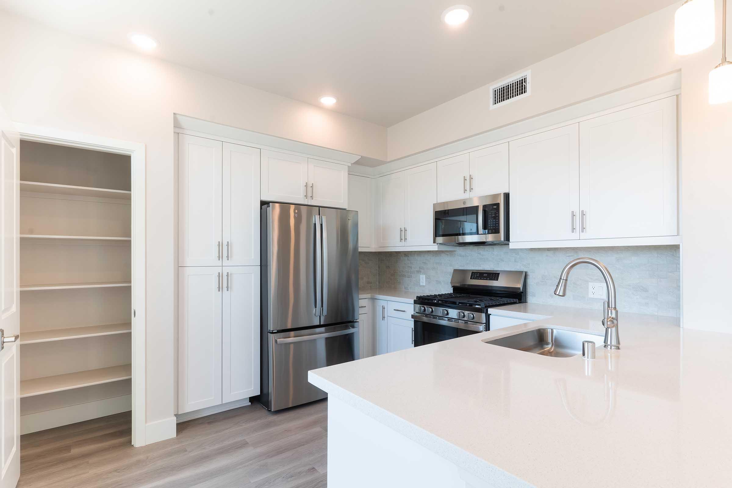 Modern kitchen featuring white cabinetry, stainless steel appliances including a refrigerator, oven, and microwave. There are quartz countertops, a single basin sink with a stylish faucet, and a spacious pantry with double doors. The lighting is bright and inviting, enhancing the contemporary design.