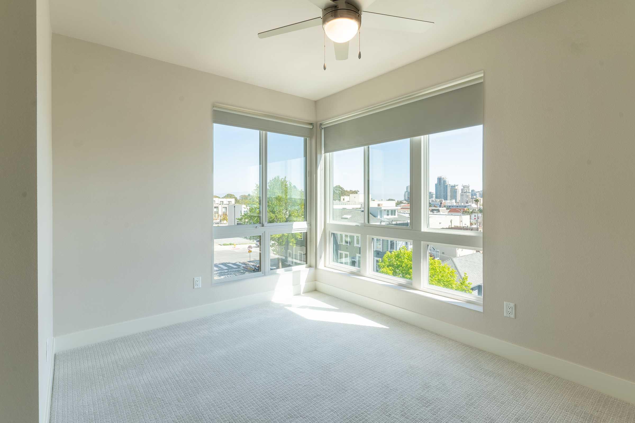 A bright, contemporary room with large windows offering a view of the city. Natural light floods the space, highlighting the neutral-colored walls and carpet. A ceiling fan is visible, adding to the modern design. The overall ambiance is open and airy, ideal for a living or bedroom area.