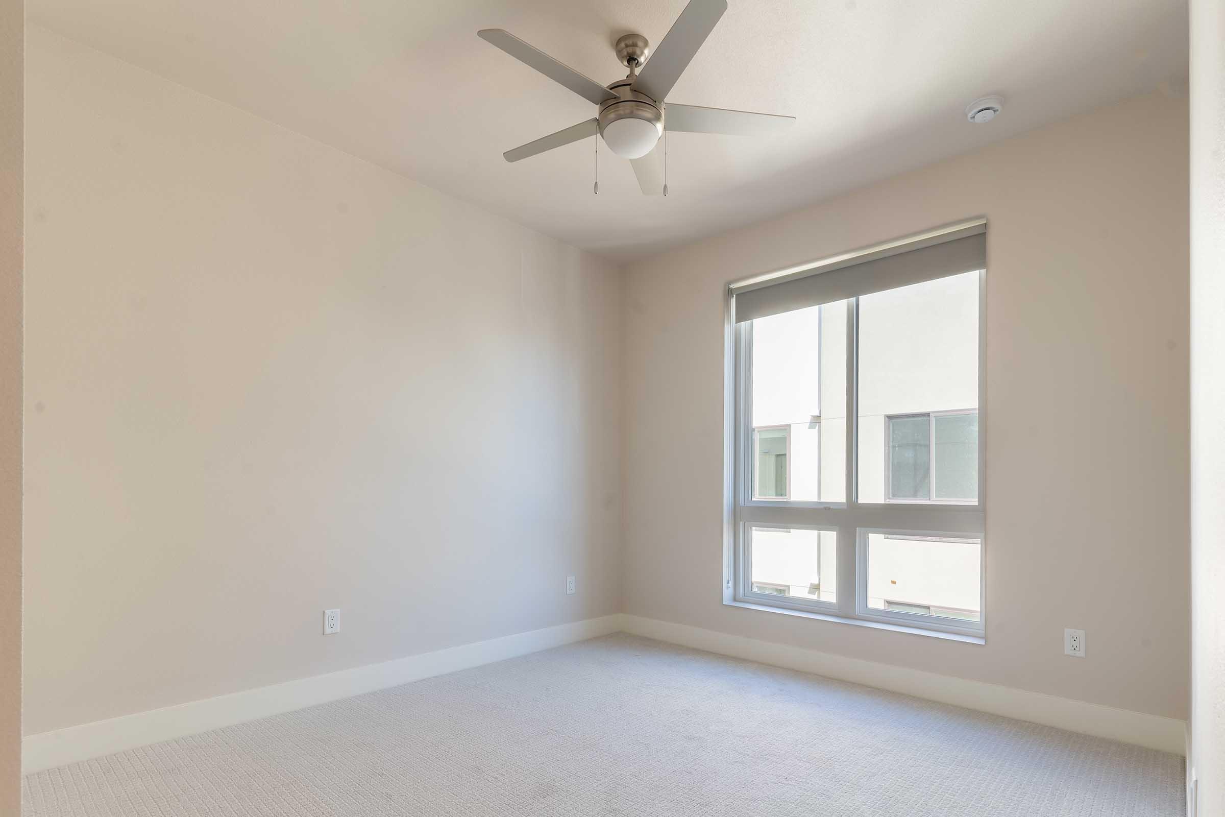 A bright, empty room featuring light-colored walls and a ceiling fan. Large windows allow natural light to enter, and the floor is covered with light carpet. The space is minimalist and ready for decoration or furniture.