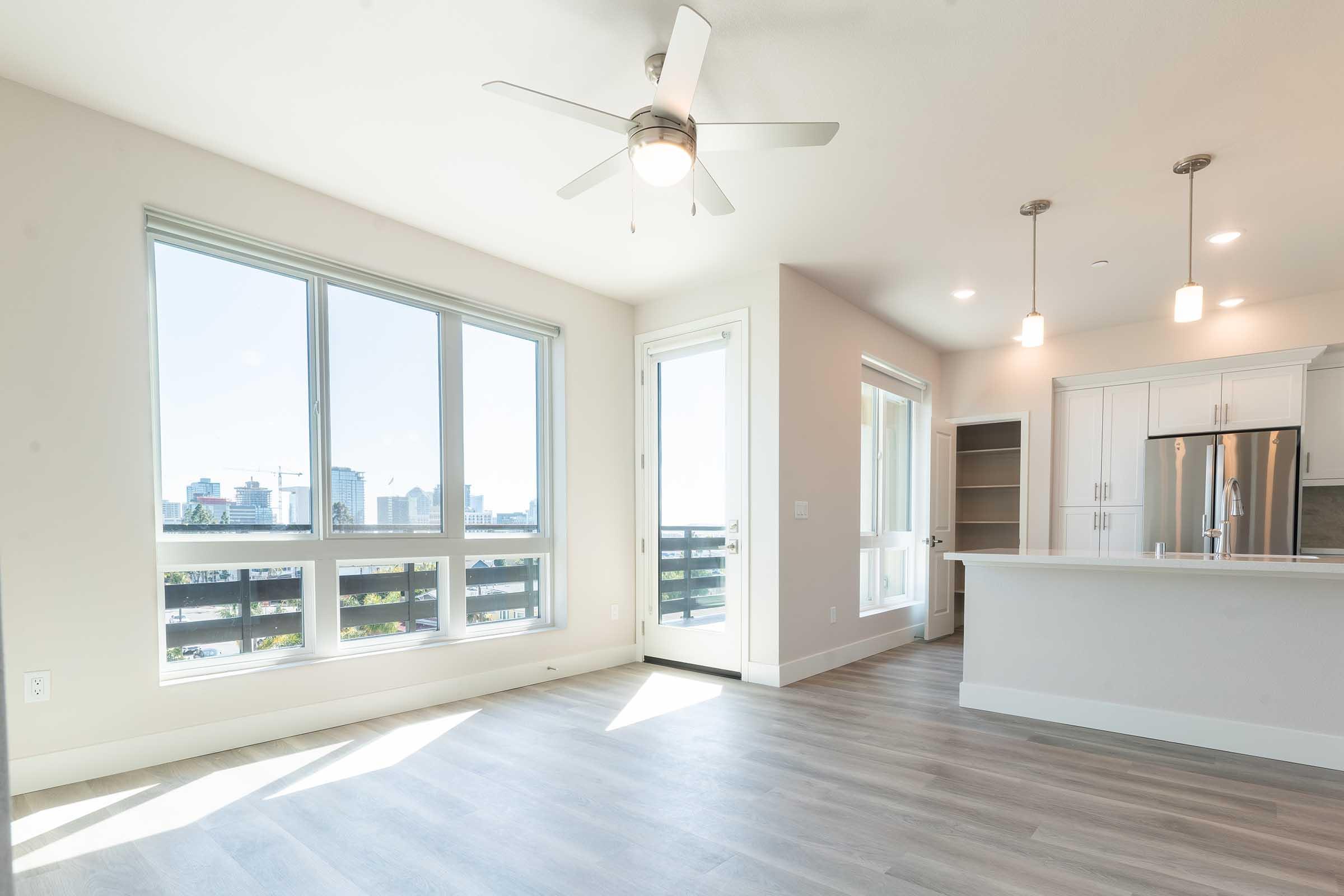 Bright and airy modern apartment interior featuring large windows with a city view, a ceiling fan, and an open kitchen area. Light-colored wood flooring complements the neutral walls. The kitchen includes white cabinetry and stainless steel appliances. A sliding glass door leads to a balcony.