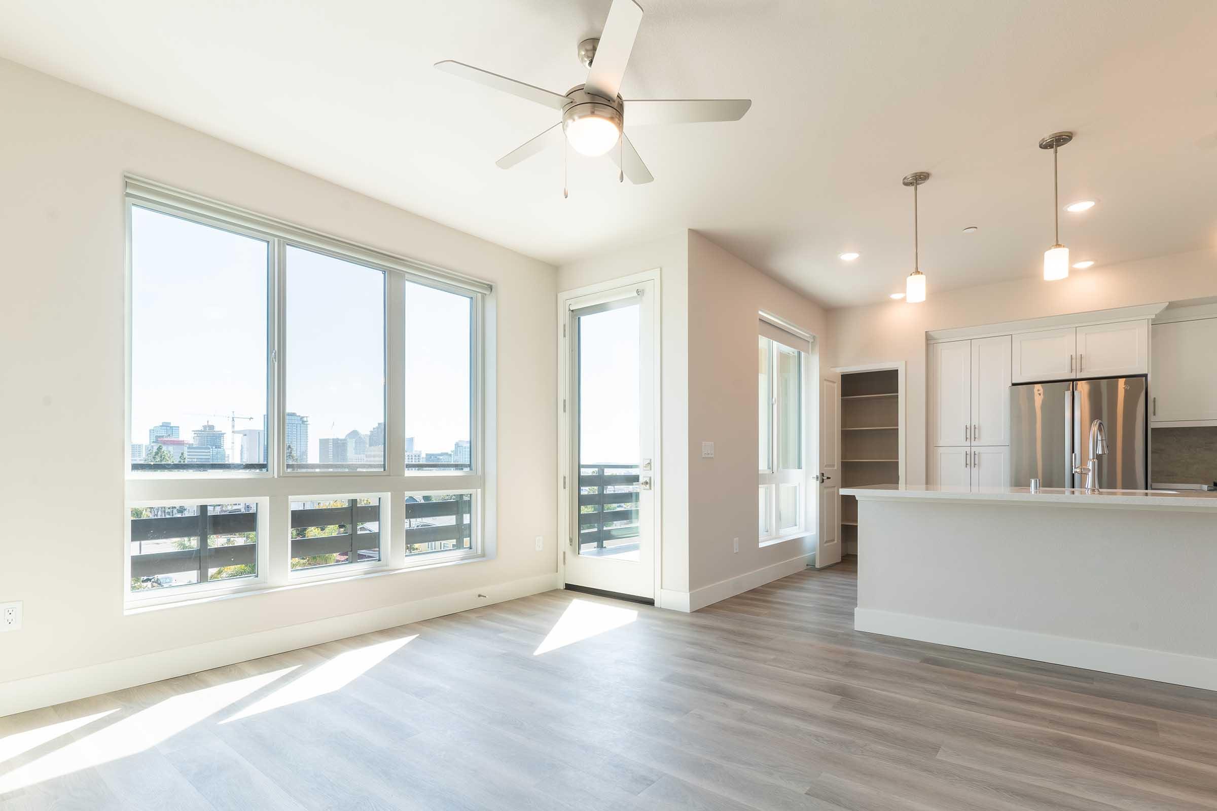 A bright and modern apartment interior featuring large windows with city views, a ceiling fan, and light wood-style flooring. The open layout includes a kitchen with white cabinets and countertop, along with a sliding door leading to a balcony. Natural light fills the space, creating a welcoming atmosphere.