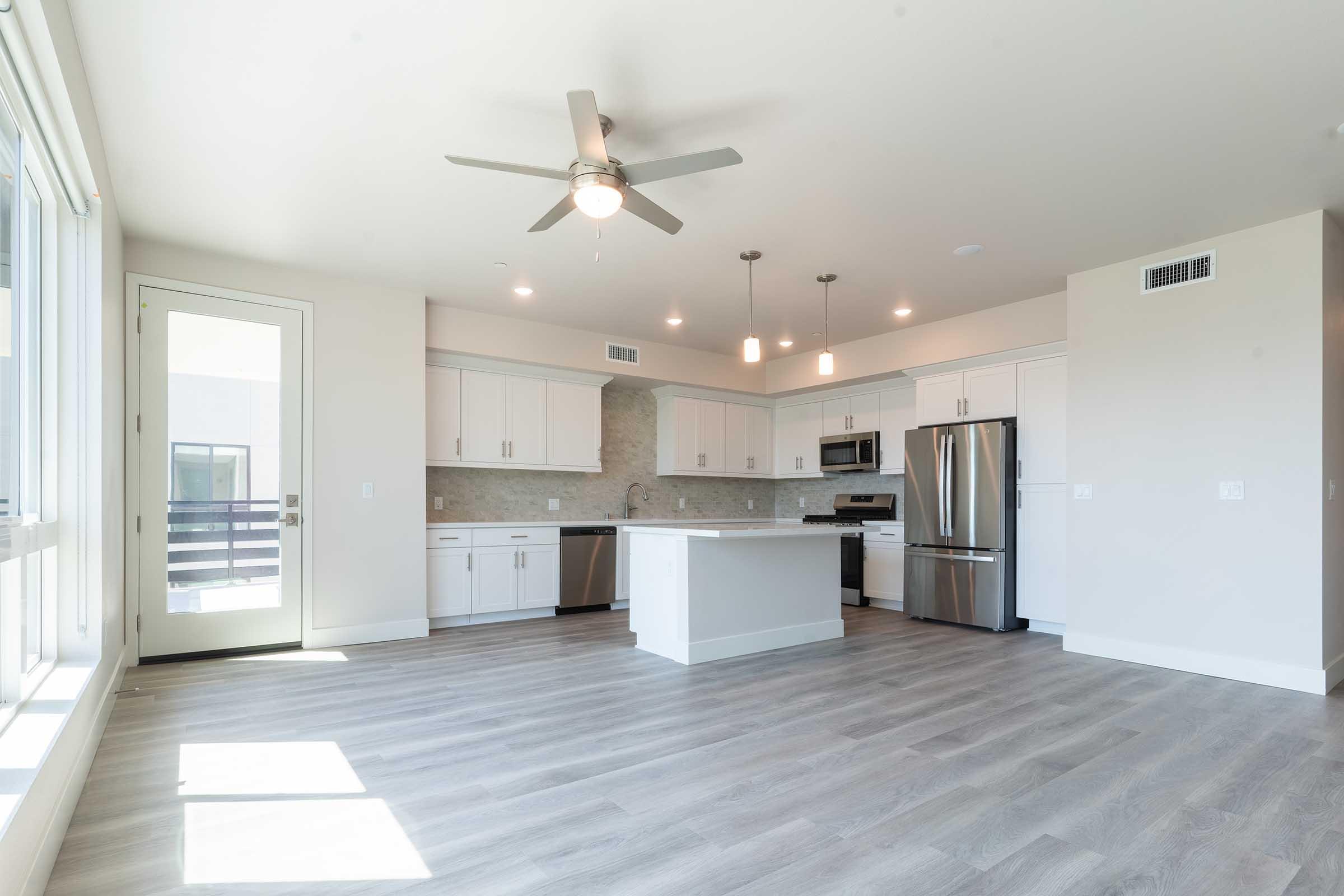 Spacious modern kitchen and living area with light-colored cabinets, stainless steel appliances, and a center island. Large windows let in natural light, and a ceiling fan provides airflow. The floor features light wood-like flooring, creating a bright and inviting atmosphere.