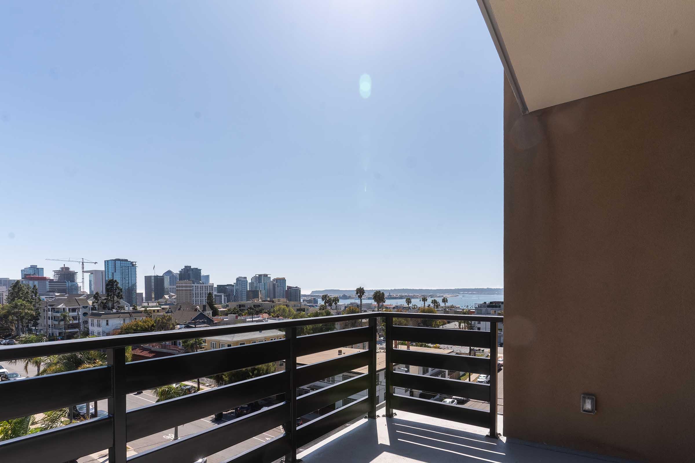 View from a balcony overlooking a city skyline with tall buildings, palm trees, and a body of water. The sky is clear and bright, indicating a sunny day. The railing of the balcony is in the foreground, framing the scenic view.