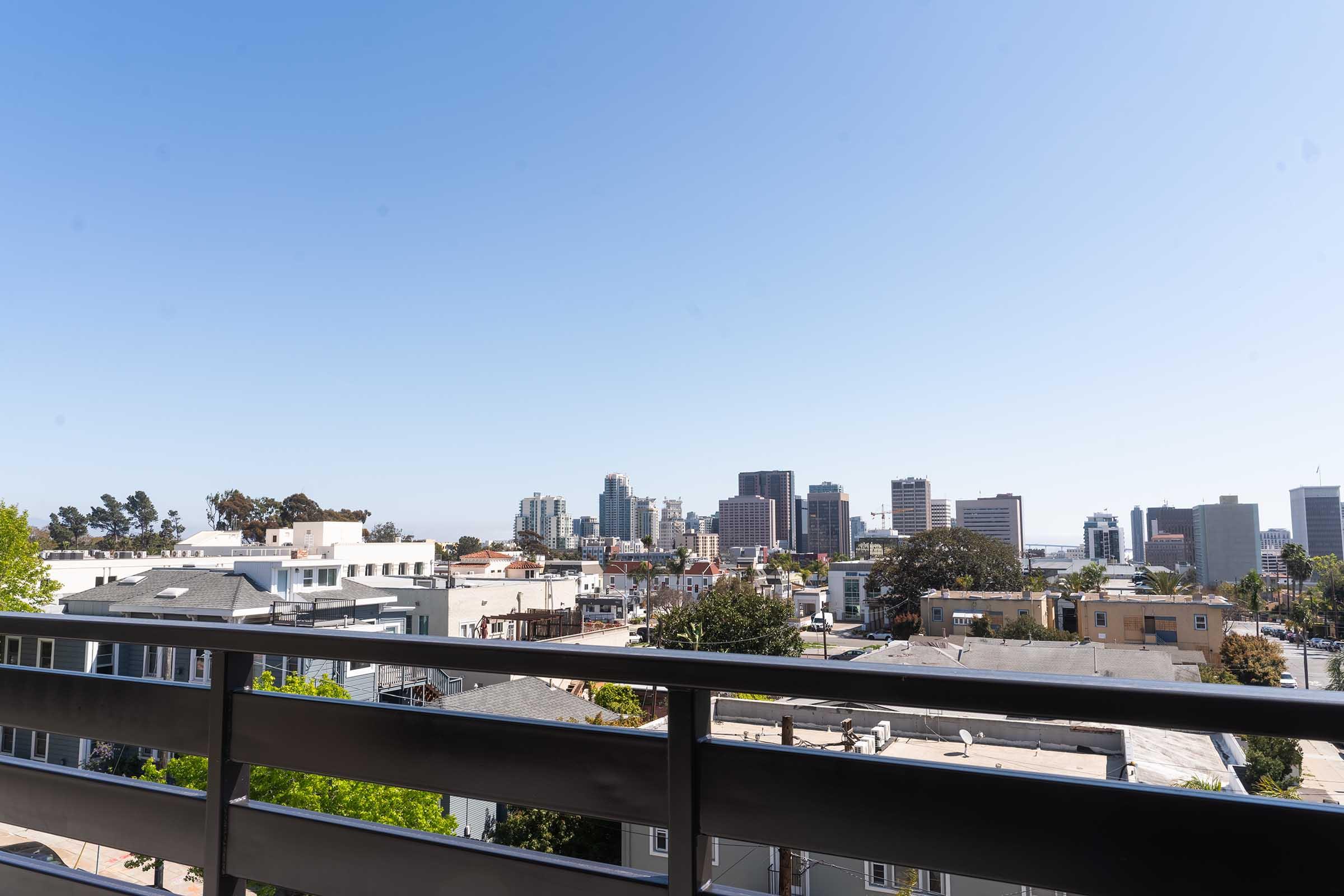 A clear blue sky over a city skyline featuring modern buildings. In the foreground, rooftops and trees are visible, with a balcony railing framing the view. The scene captures a sunny day in an urban environment, showcasing a blend of residential and commercial architecture.