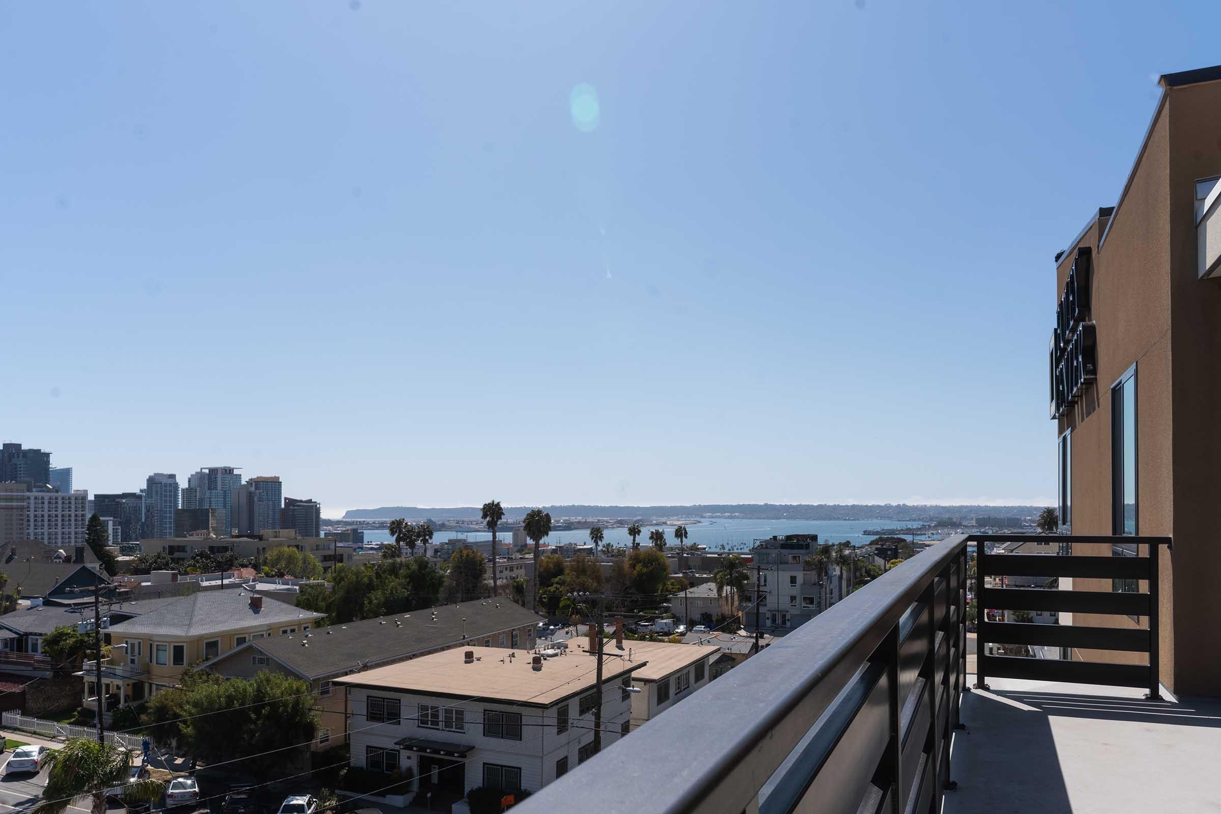 View from a balcony overlooking a cityscape, featuring modern buildings, palm trees, and a glimpse of the water in the distance under a clear blue sky. The foreground includes rooftops and streets, while the background shows urban architecture along the waterfront.