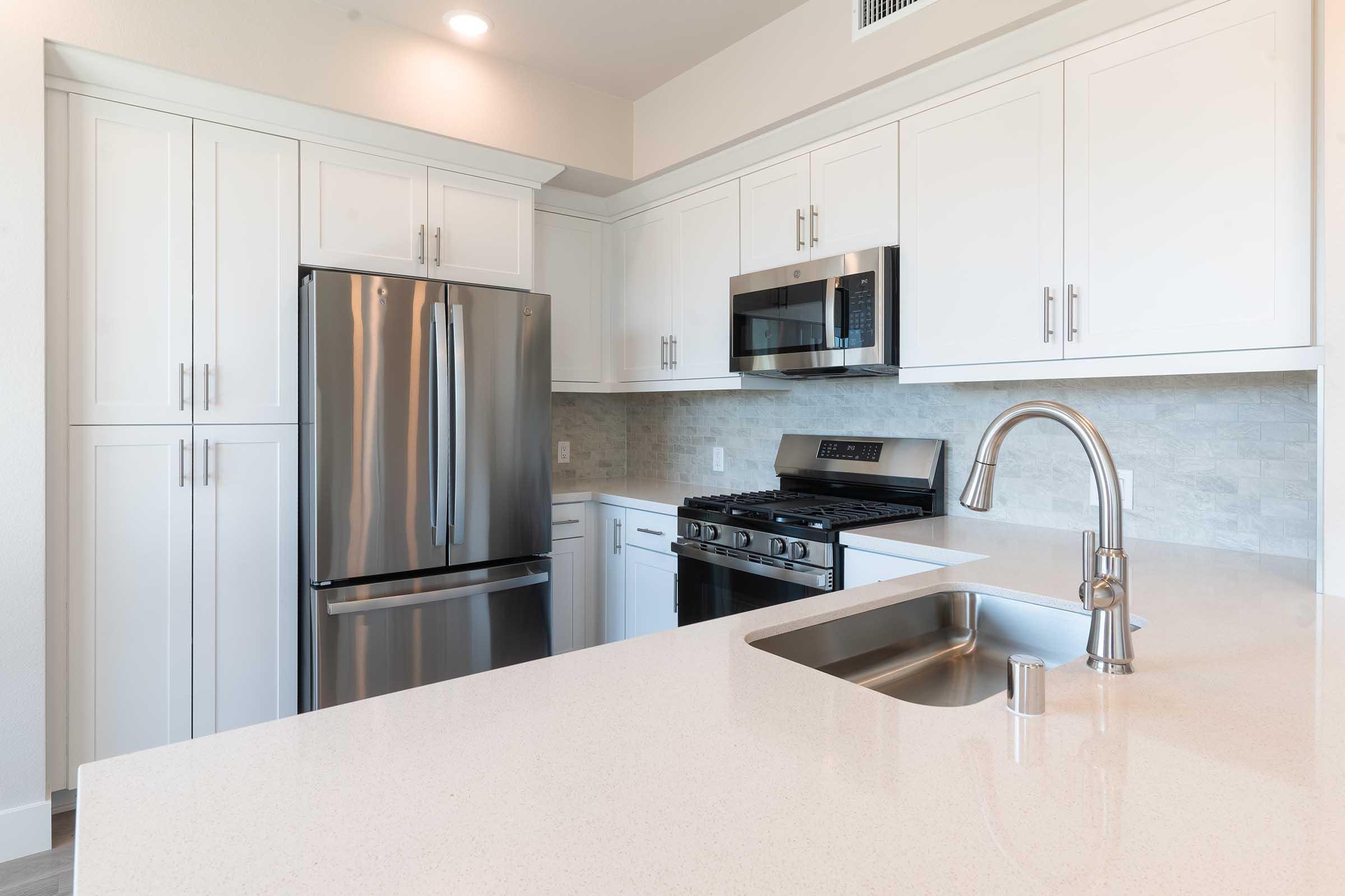 A modern kitchen featuring white cabinetry, a stainless steel refrigerator, microwave, and gas stove. The countertop is a light-colored stone with a sink and faucet. The backsplash is a subtle textured finish, creating a clean and contemporary aesthetic. Natural light brightens the space.