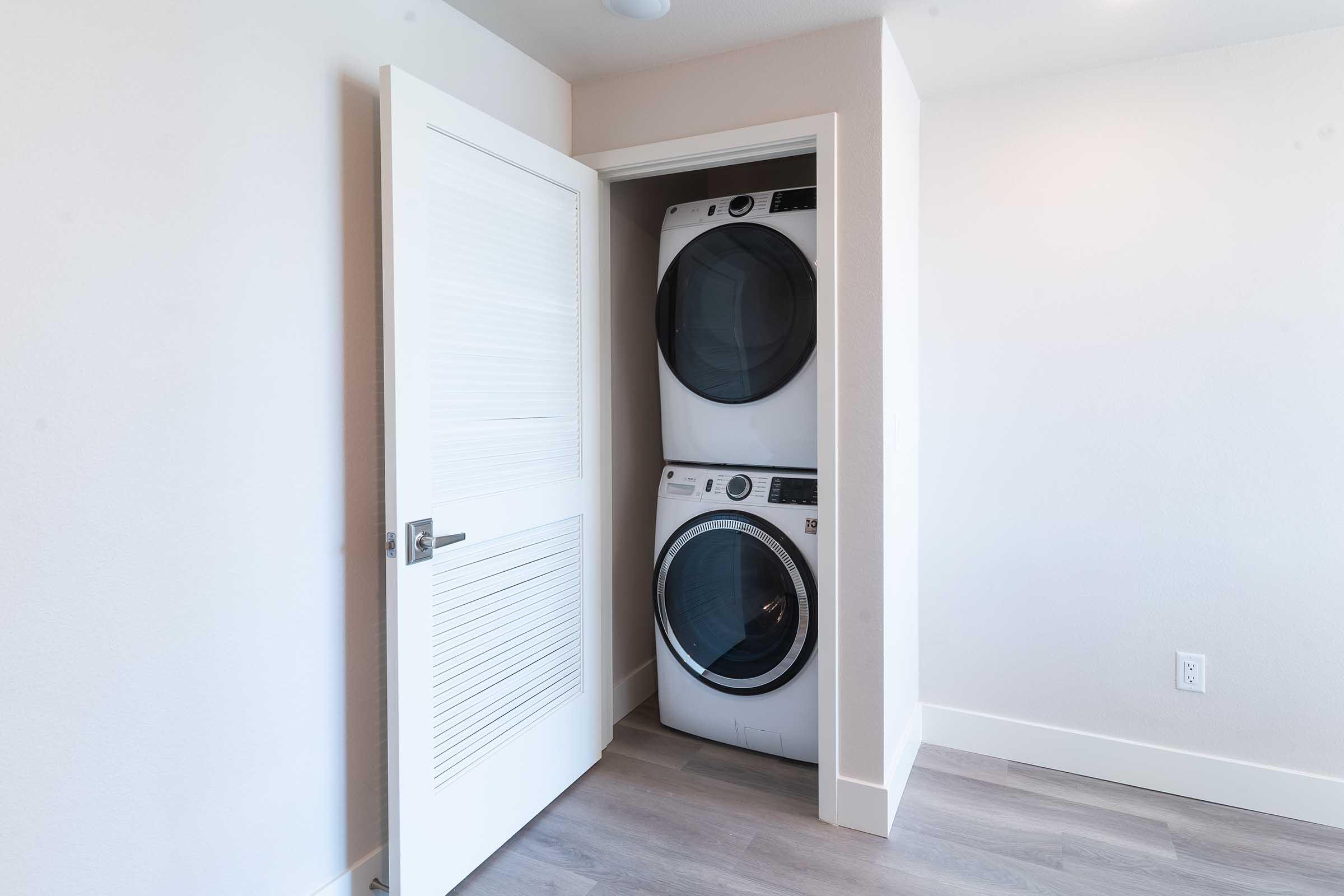A compact laundry space featuring a stacked washer and dryer within a white closet. The door is open, revealing the appliances against a light-colored wall and modern flooring. The room has a clean, minimalistic design with ample natural light.