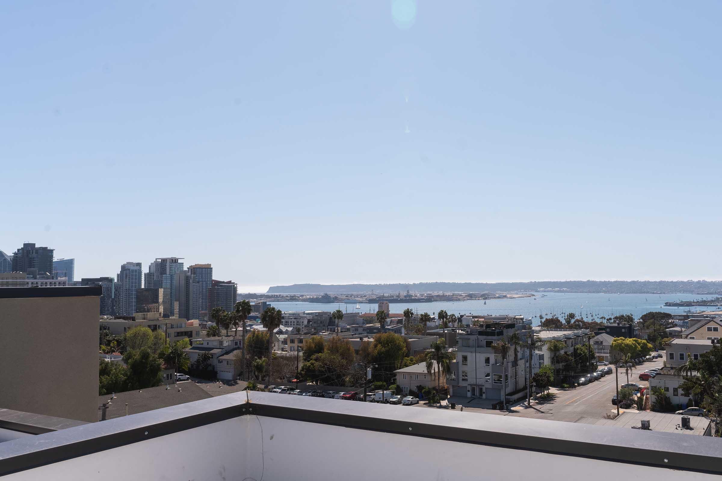 A panoramic view of a coastal city featuring modern buildings, palm trees, and a glimpse of the water from a rooftop terrace. The clear blue sky complements the urban landscape, with distant land visible across the water.