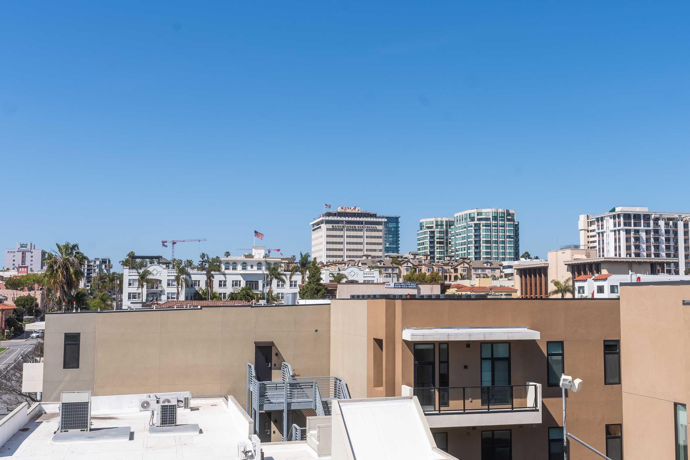 A view of a city skyline featuring various buildings under a clear blue sky. In the foreground, rooftops of modern structures are visible, with palm trees and construction cranes hinting at urban development. The background showcases high-rise buildings, including a hotel and residential complexes.