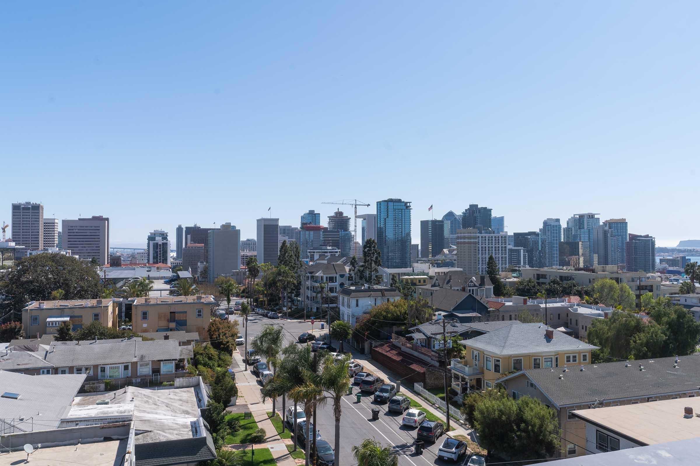 A panoramic view of a city skyline featuring modern skyscrapers and a mix of residential buildings. In the foreground, a neighborhood with palm trees and streets is visible, while the background showcases urban development with clear blue skies and distant cranes indicating construction.
