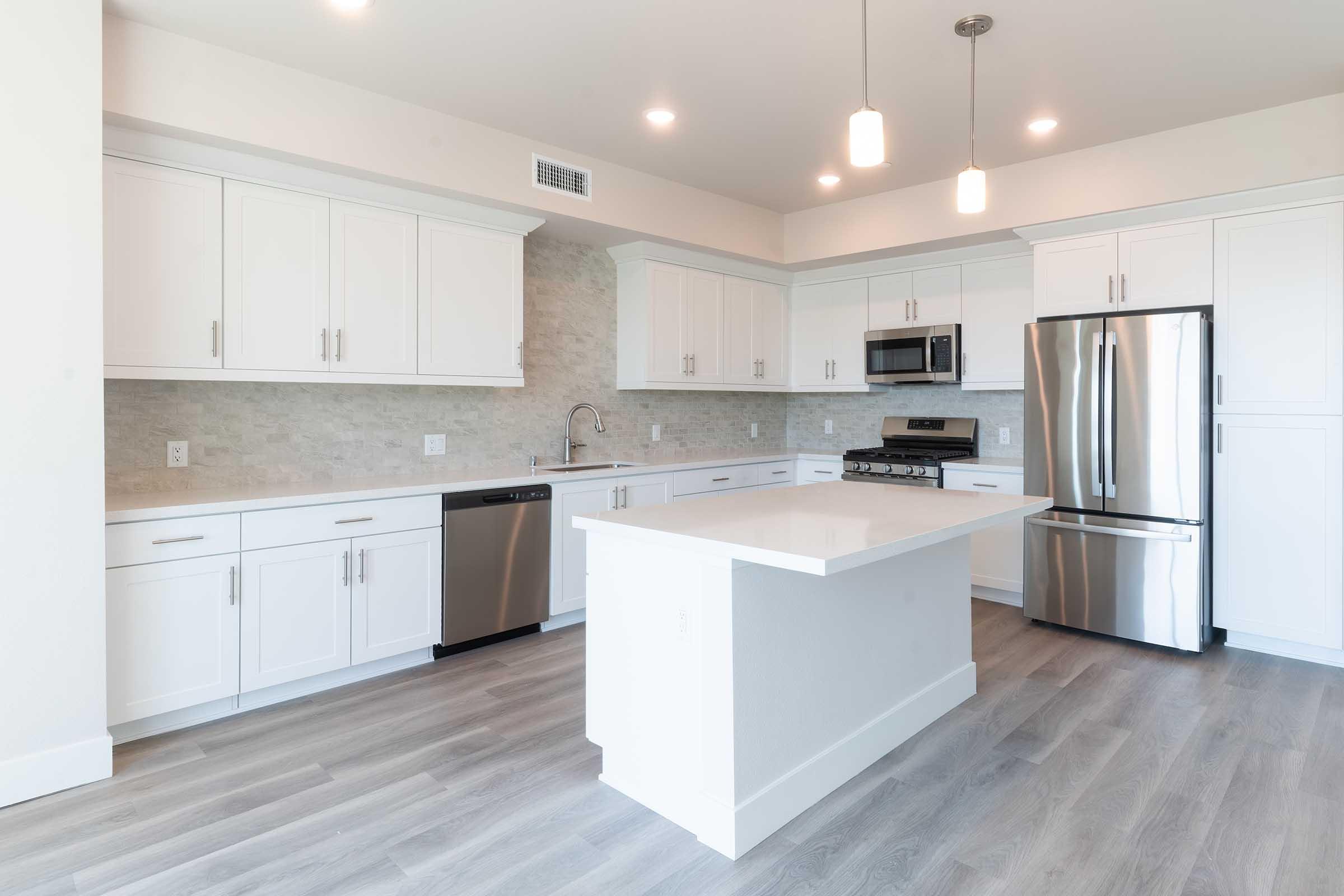 Modern kitchen with white cabinetry, stainless steel appliances, a large island with a countertop, and a light gray backsplash. Bright lighting fixtures hang above the island, and the floor features light gray wood-like tiles. The overall design is clean and spacious, ideal for a contemporary home.