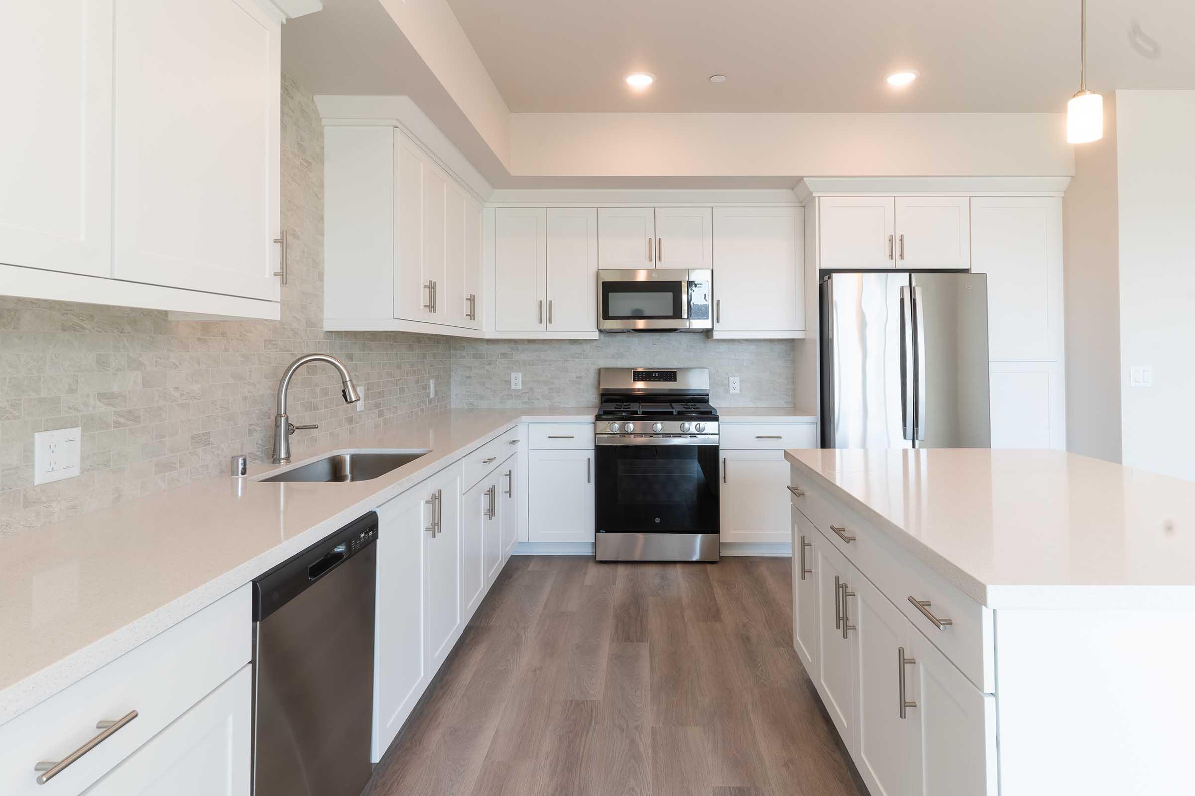 Modern kitchen featuring white cabinetry, stainless steel appliances including a microwave and gas stove, and a spacious island with a pristine quartz countertop. The design includes a stylish backsplash and wood-like flooring, creating a bright and open atmosphere. Natural light floods the space from large windows.