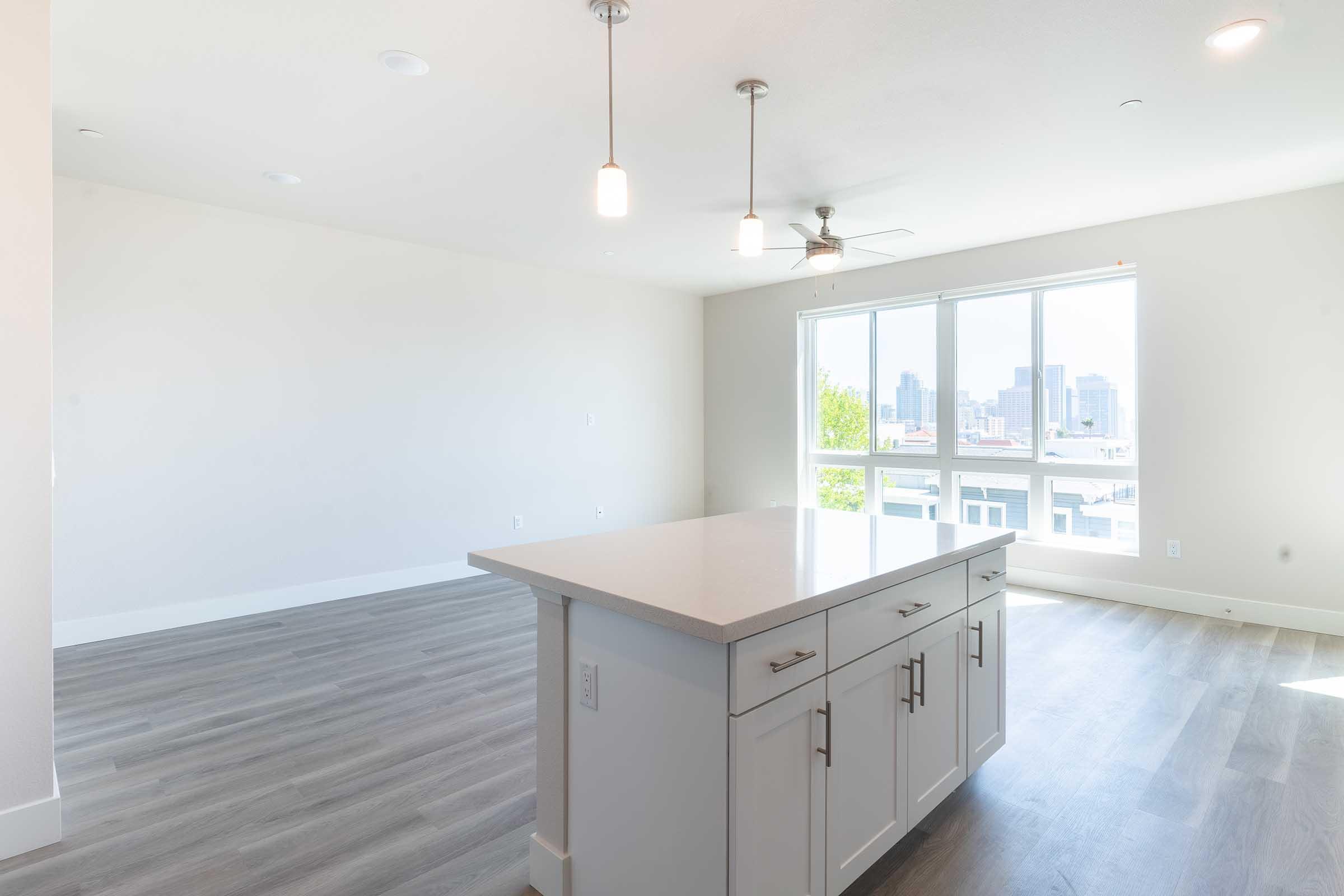 Bright and airy modern living space featuring a kitchen island with a white countertop, pendant lights overhead, and large windows with a city view. The floor is covered with light gray laminate, and the walls are painted in neutral tones, creating an open and inviting atmosphere.