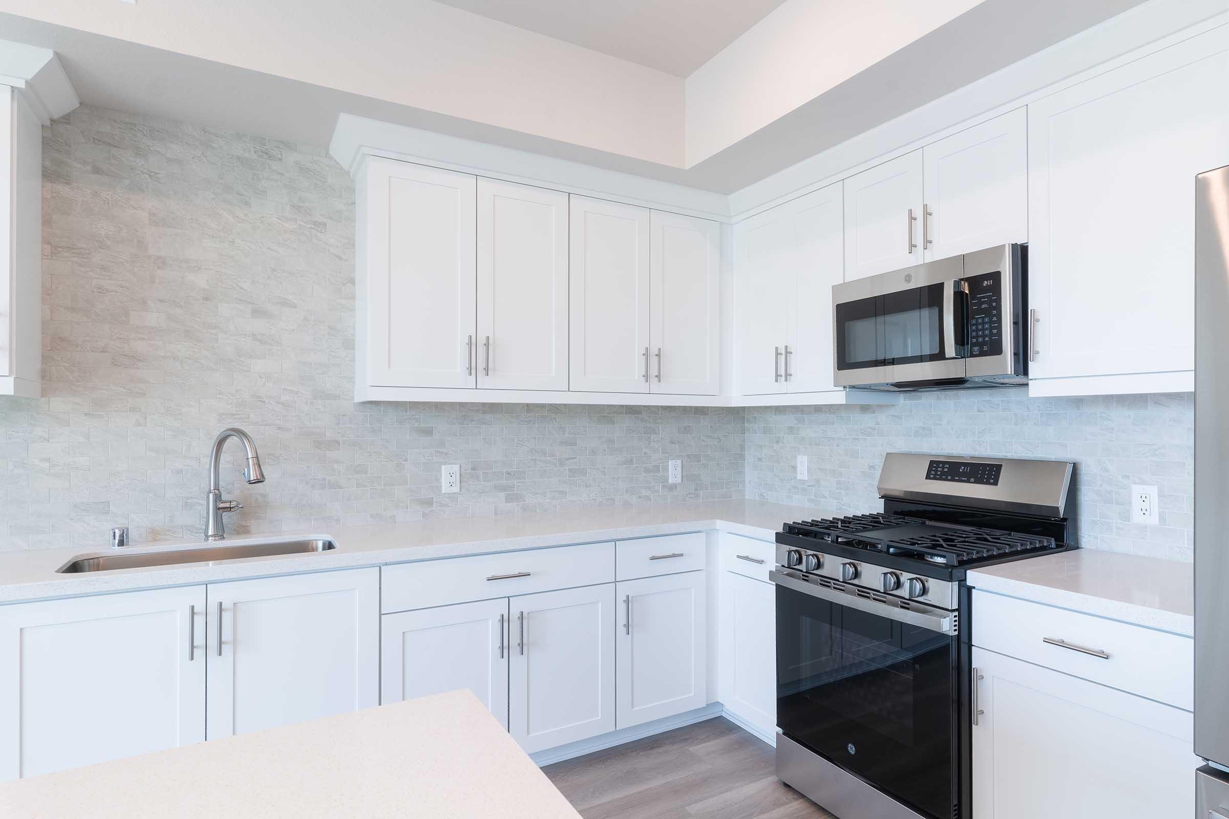 Modern kitchen with white cabinetry, stainless steel appliances including a microwave and gas stove, and a sleek sink. The backsplash features light-colored tiles, and the countertops are a smooth, neutral shade. The space has ample natural light, creating a clean and airy atmosphere.