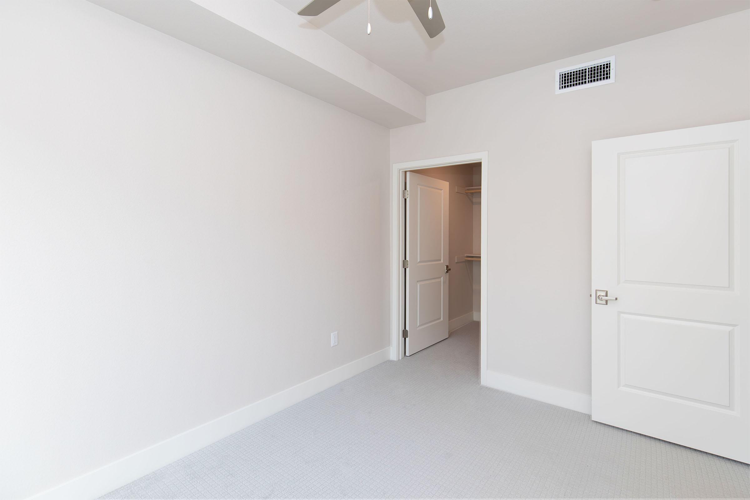 Empty bedroom featuring light-colored walls, a ceiling fan, and a closet visible through an open door. The carpeted floor is neutral in tone, and the space is well-lit with natural light. The overall appearance is clean and minimalistic, ideal for customization.