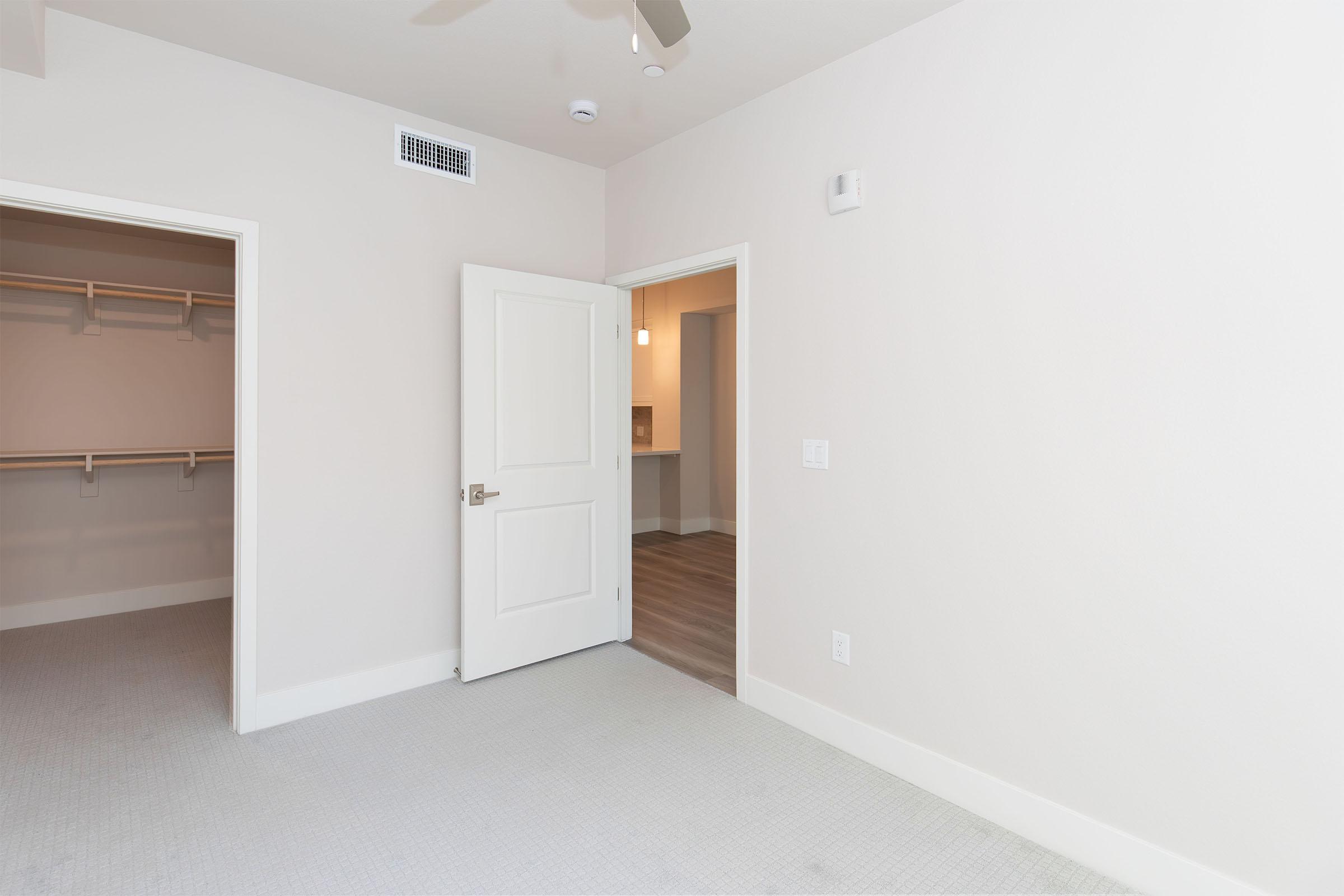 A light-colored room featuring an open door leading to another space. There is a closet with a hanging rack on the left side, and the floor is carpeted. The walls are smooth and unadorned, creating a clean, minimal look. Natural light can be seen coming from the adjacent room through the open doorway.