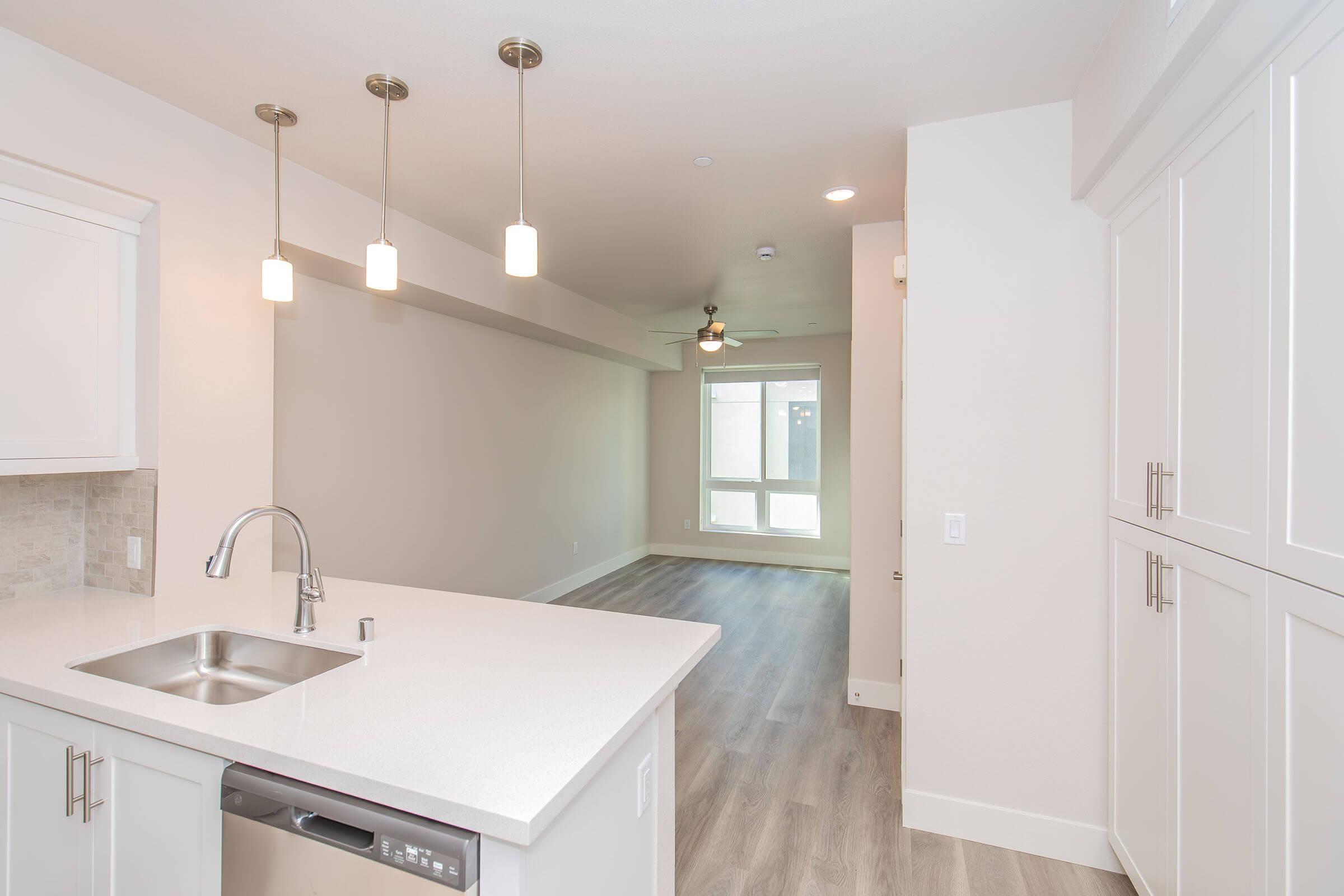 Modern kitchen with a white countertop and cabinet, featuring a stainless steel sink. In the background, a spacious living area with light-colored walls, large windows allowing natural light, and light wood flooring. Pendant lights hang from the ceiling, adding a contemporary touch.