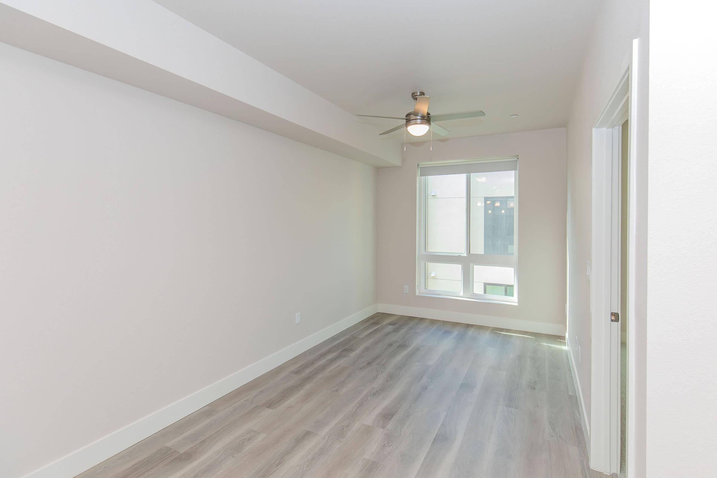 A minimalistic, empty room with light-colored walls and a ceiling fan. A window allows natural light to enter, illuminating the wooden floor. The space has a modern feel, ideal for various uses such as a living room or office. A door on the right leads to another area.