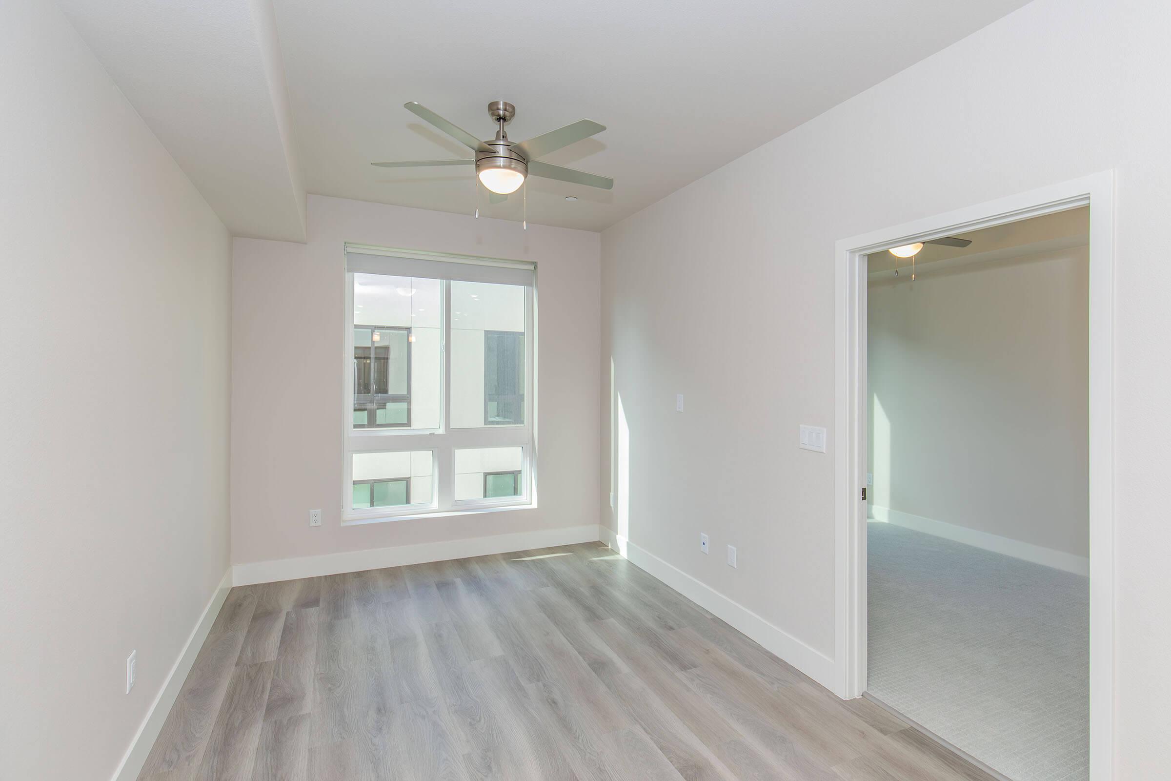 A bright, empty room featuring light-colored walls and a ceiling fan. A large window allows natural light to enter, and there's a door leading to another room. The flooring is a light wood, creating a clean and spacious appearance.