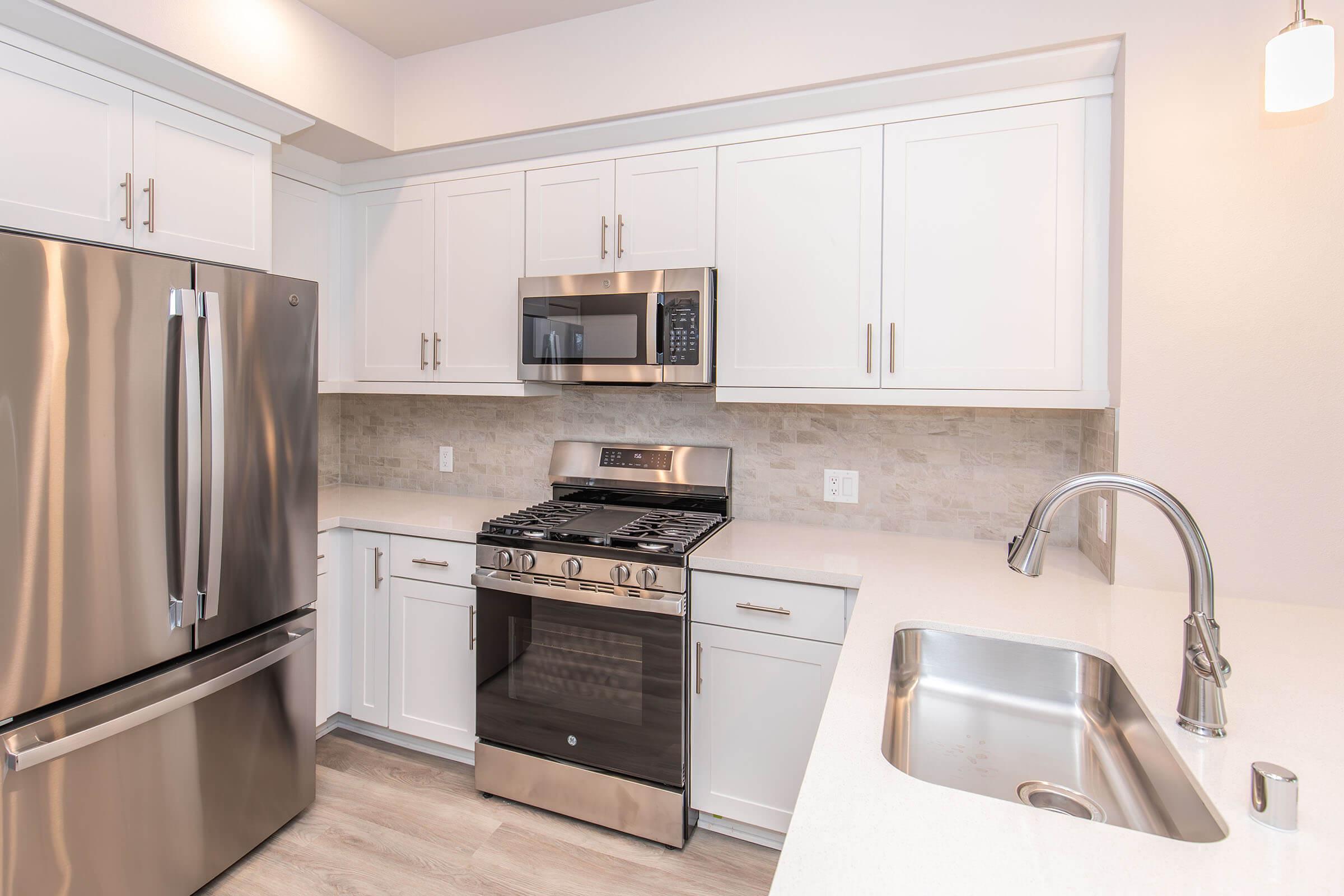 Modern kitchen featuring stainless steel appliances, including a refrigerator, microwave, and gas stove. The sleek cabinetry is white with a light-colored backsplash, and there’s a stainless steel sink with a modern faucet. The floor is light wood, creating a bright and contemporary atmosphere.