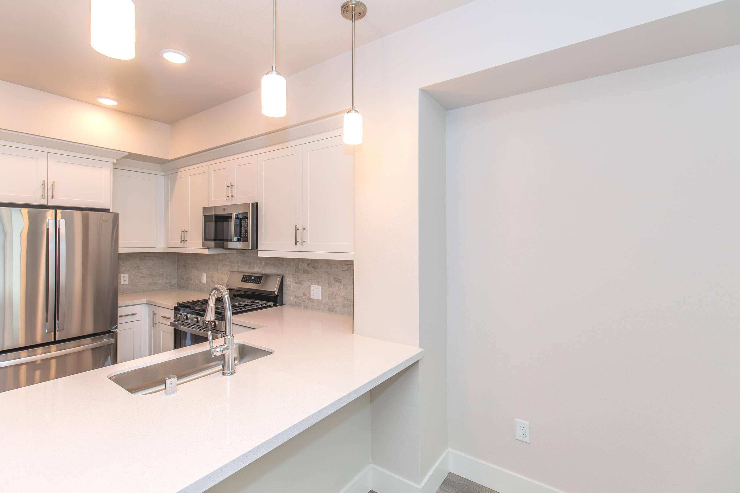 Modern kitchen featuring white cabinetry, a stainless steel refrigerator, and a gas stove. The countertop is sleek and white with a double basin sink. There are pendant lights hanging above the island, and the walls are painted in a light color, enhancing the bright and open feel of the space.