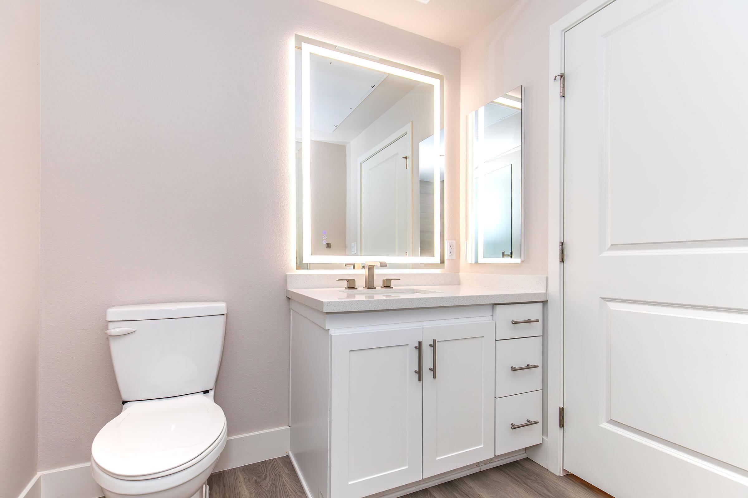 A modern bathroom featuring a white cabinet with a sink, a large illuminated mirror, and a white toilet. The walls are painted in a light color, and the flooring has a wood-like finish. There are two windows providing natural light, enhancing the contemporary aesthetic.
