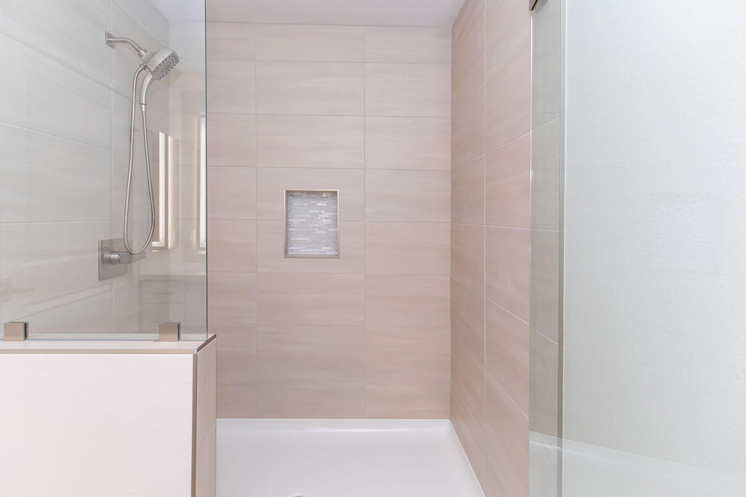 A modern shower stall with light-colored tiled walls and a glass door. The space features a chrome showerhead mounted on the wall and a built-in niche for holding toiletries. The shower base is white, creating a clean and contemporary look.