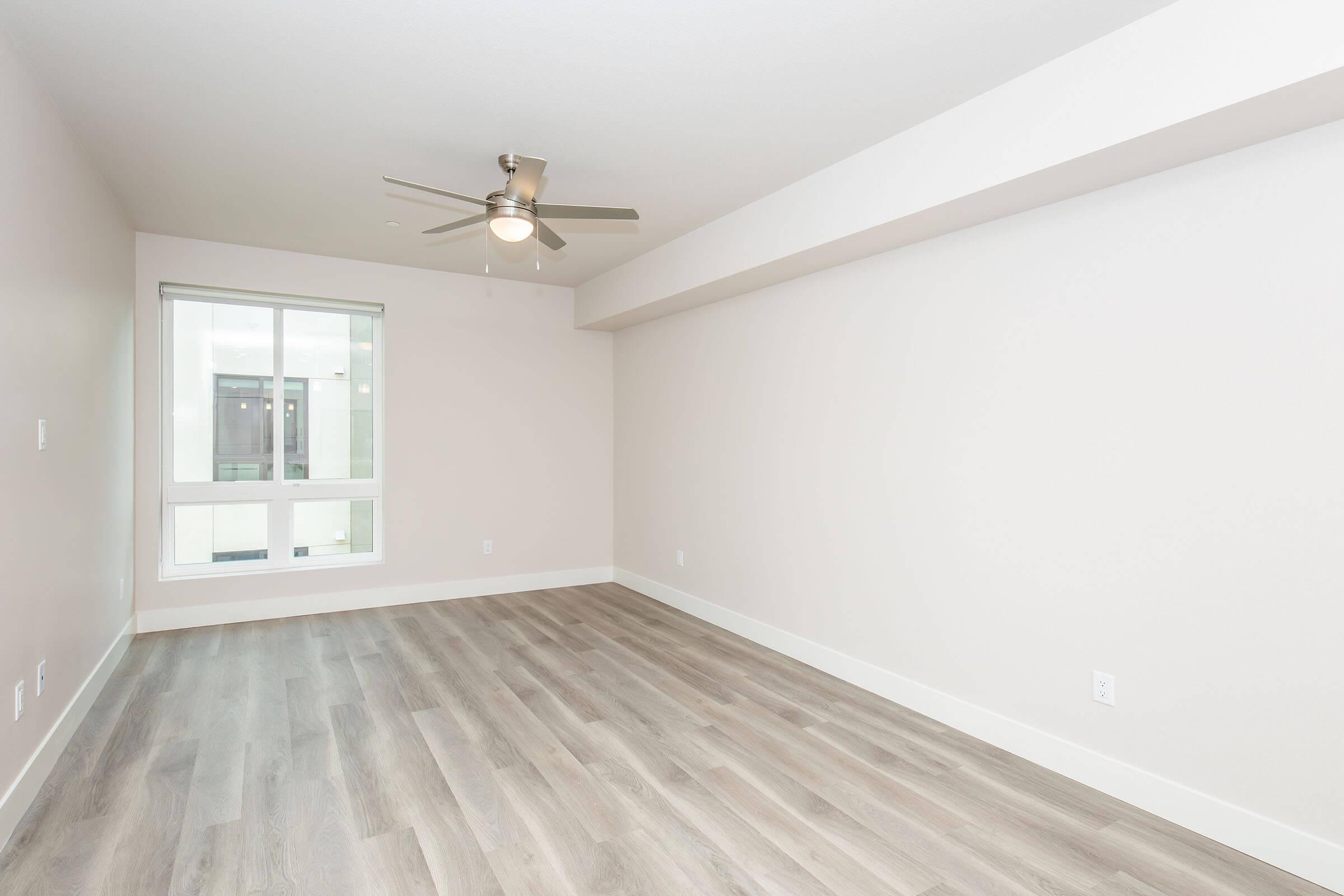 Empty room with light-colored walls and wood-look flooring. A ceiling fan is mounted in the center. A large window lets in natural light, revealing a view outside. The space is unfurnished, giving it a clean and modern appearance.