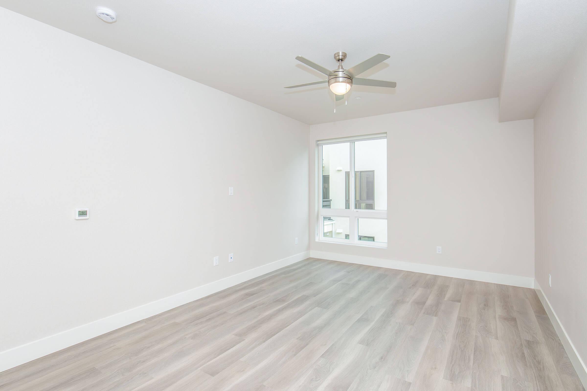 A bright, empty room with light-colored walls and wood-like flooring. A ceiling fan is mounted in the center, and a large window lets in natural light. The space appears clean and modern, suitable for various furniture arrangements.