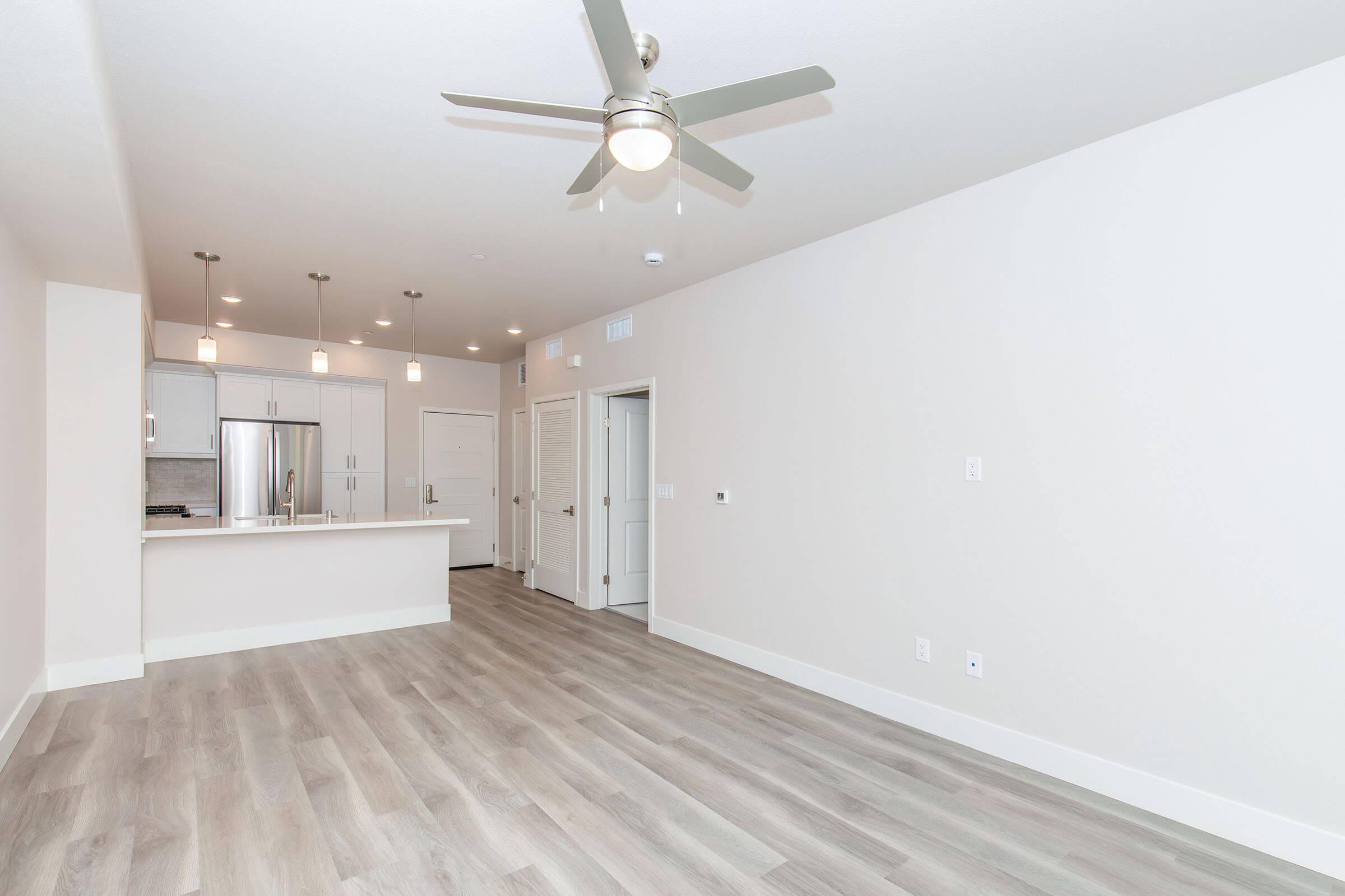 A modern, empty living room featuring light-colored walls, hardwood flooring, and a ceiling fan. The space includes an open kitchen area with pendant lighting, a refrigerator, and a doorway leading to another room. Natural light streams in through the windows, creating a bright and airy atmosphere.