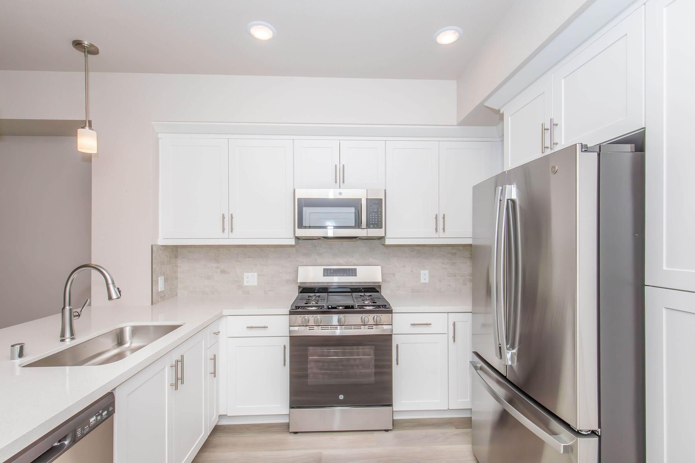 Modern kitchen featuring white cabinetry, stainless steel appliances including a gas stove and microwave, and a large double sink. The countertop is a light-colored stone, with a stylish pendant light above the sink and a spacious layout. The overall design is bright and contemporary.