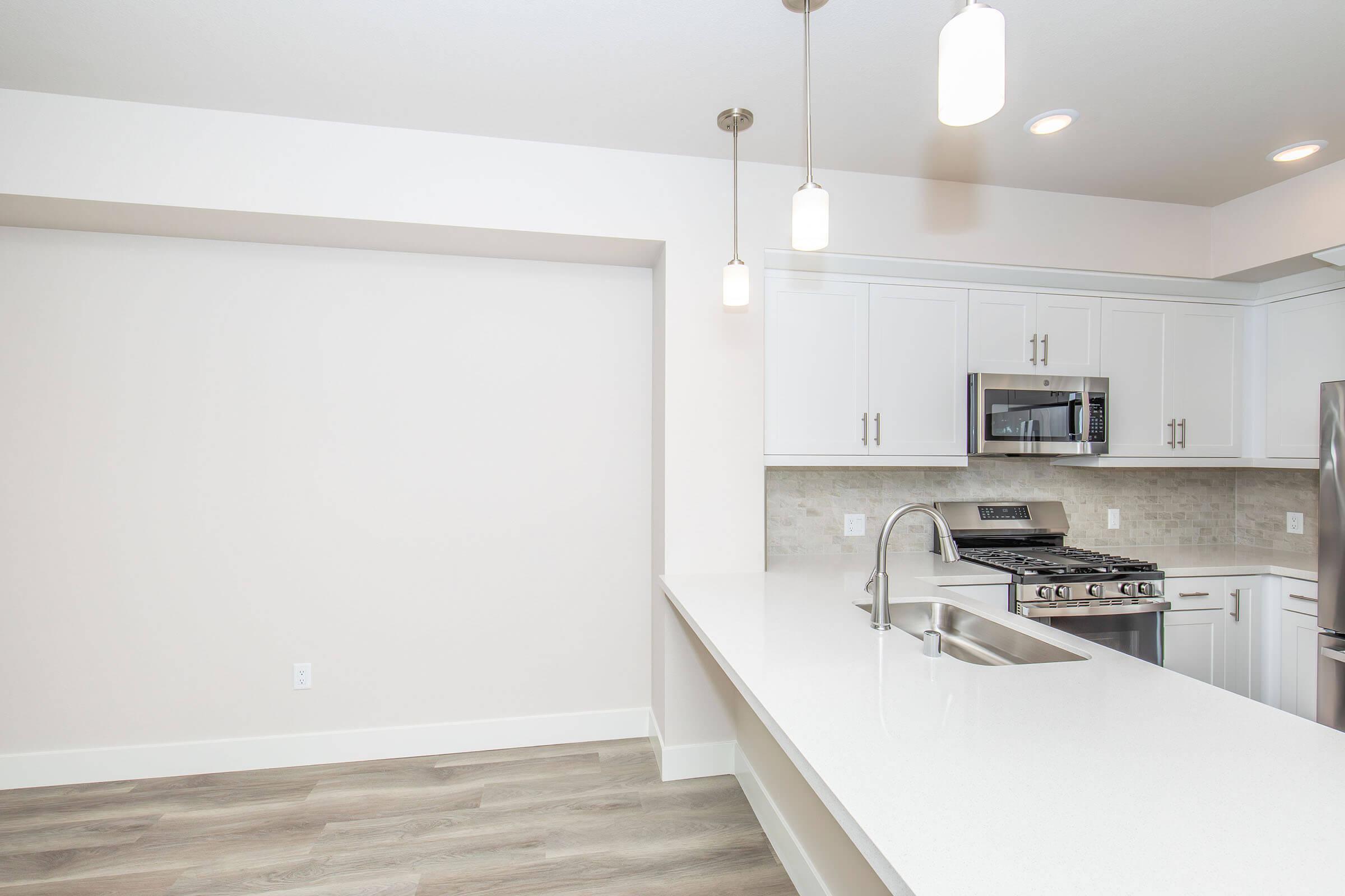 Modern kitchen with white cabinetry and stainless steel appliances. A long, white countertop extends into a spacious area with pendant lighting. The backsplash is light-colored tile, and the flooring is a warm wood tone. There is an empty wall area, providing a clean and open feel.