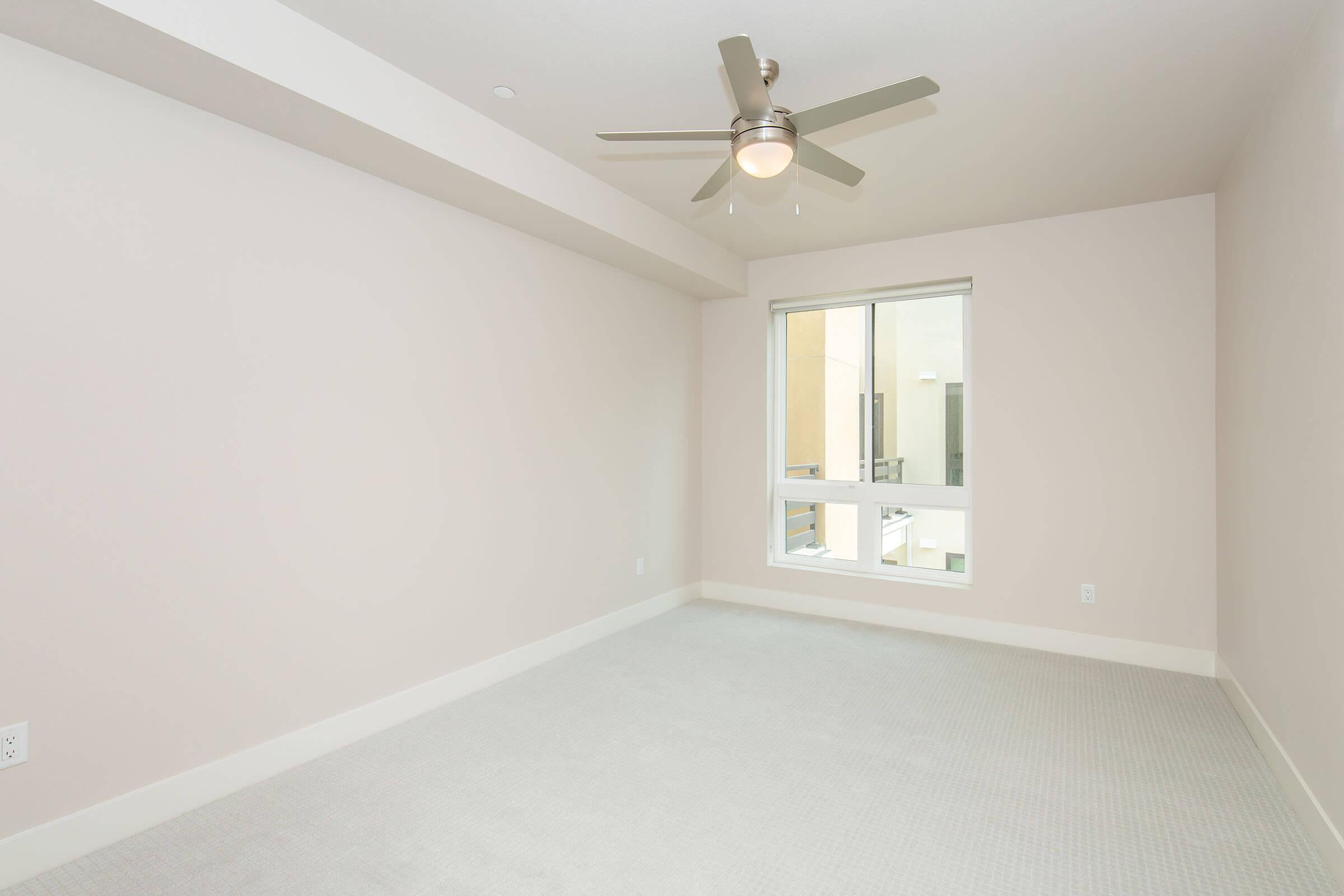 A spacious, empty room with light beige walls and a light-colored carpet. A ceiling fan is mounted on the ceiling, and there is a large window allowing natural light to enter, showing the outdoor area. The overall atmosphere is bright and airy, perfect for personalization or moving in.