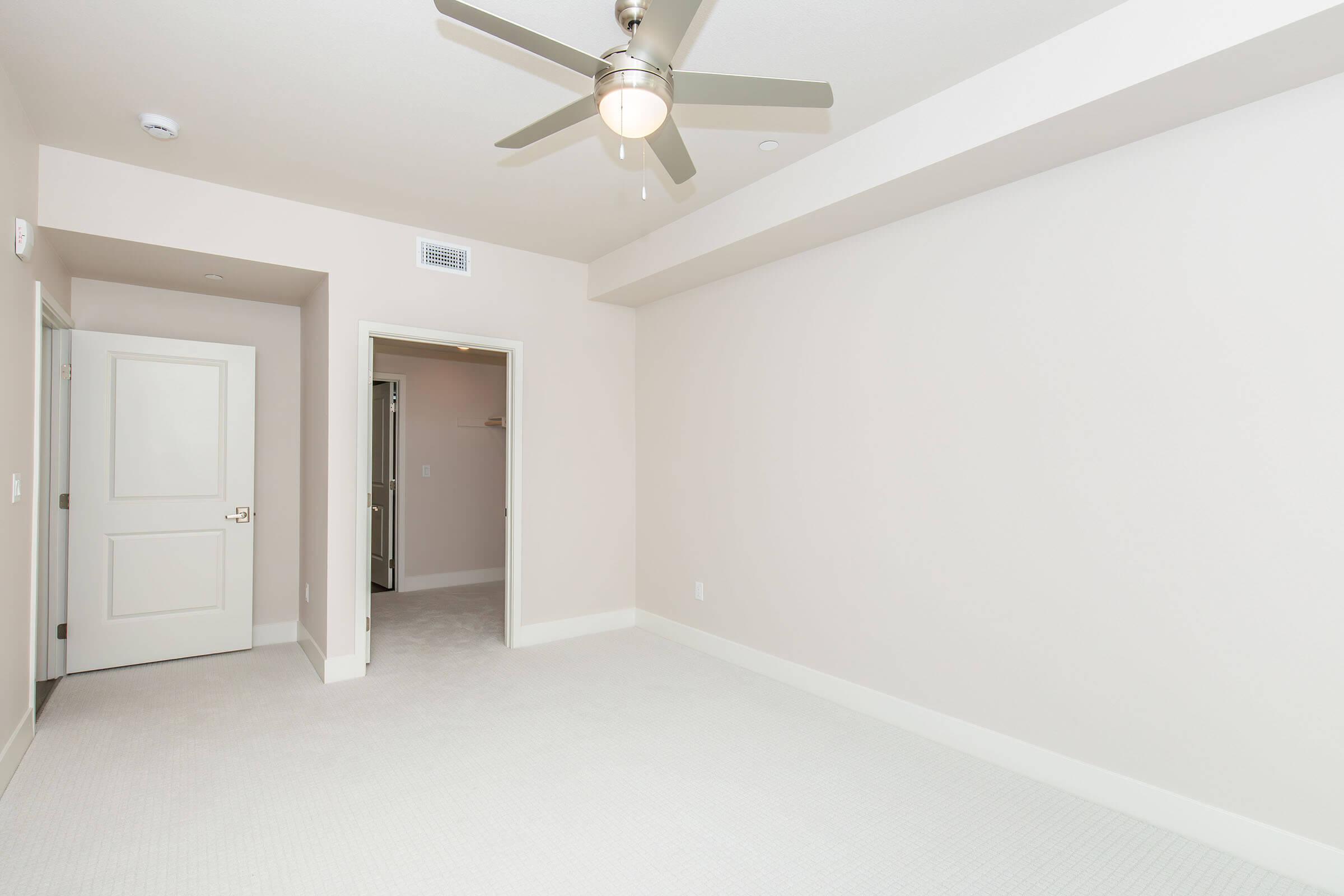 A spacious, empty room with light-colored walls and carpet. There is a ceiling fan and a doorway leading to a closet. Another door is visible on the left, leading to an adjacent room or hallway. Natural light fills the space, creating a bright and airy atmosphere.