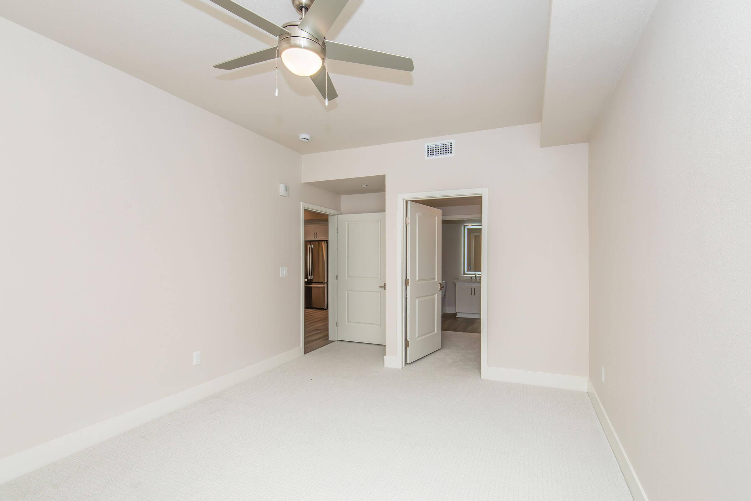 A spacious, empty room with light-colored walls and carpet. It features a ceiling fan and two doorways, one leading to a bathroom area. Natural light enters from an unseen window, creating a bright and airy atmosphere. The overall aesthetic is clean and modern, offering potential for personalization.