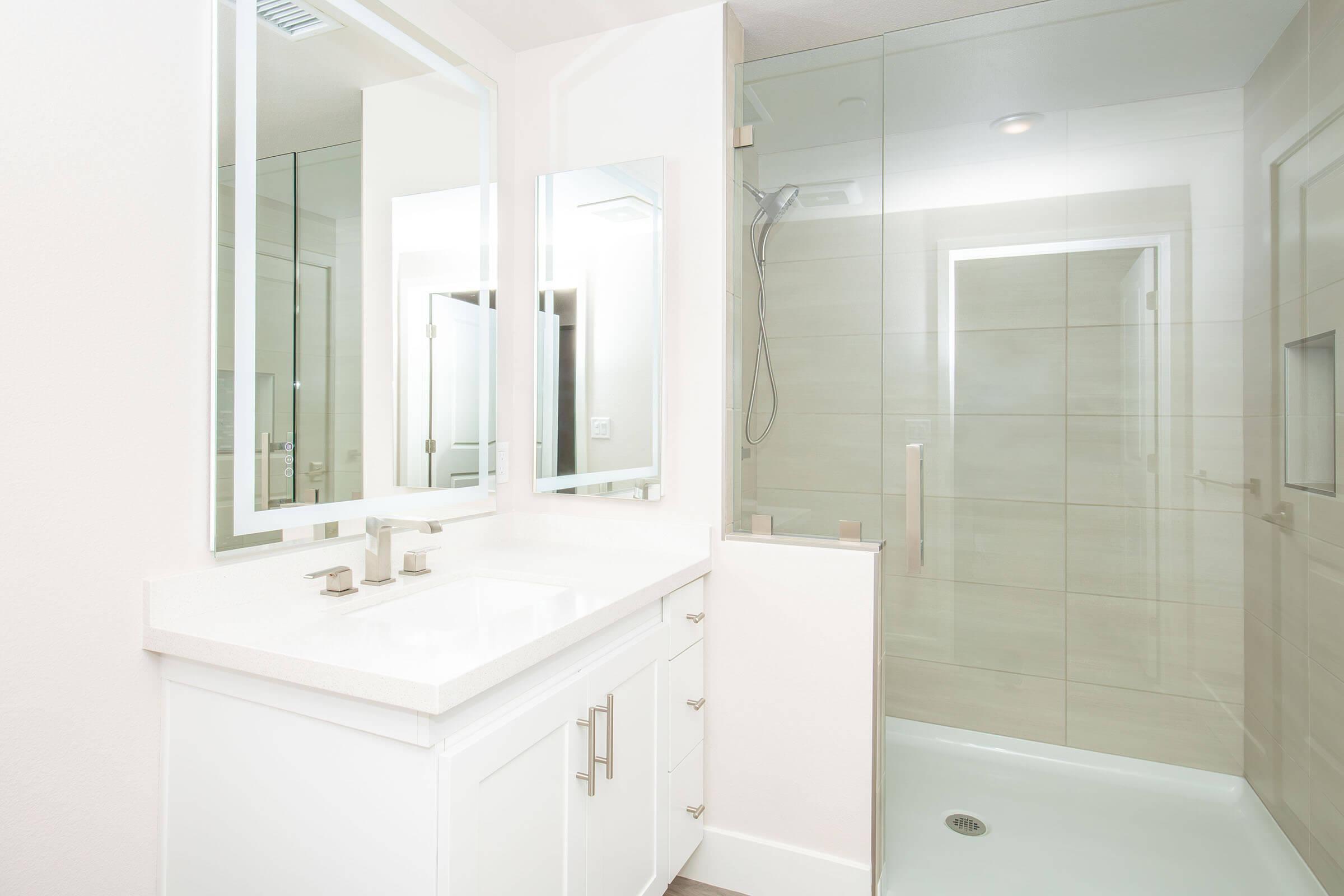 Modern bathroom featuring a white vanity with dual sinks and a large mirror. The shower area has glass doors and tiled walls, offering a clean and sleek design. Soft lighting enhances the bright, airy atmosphere of the space.
