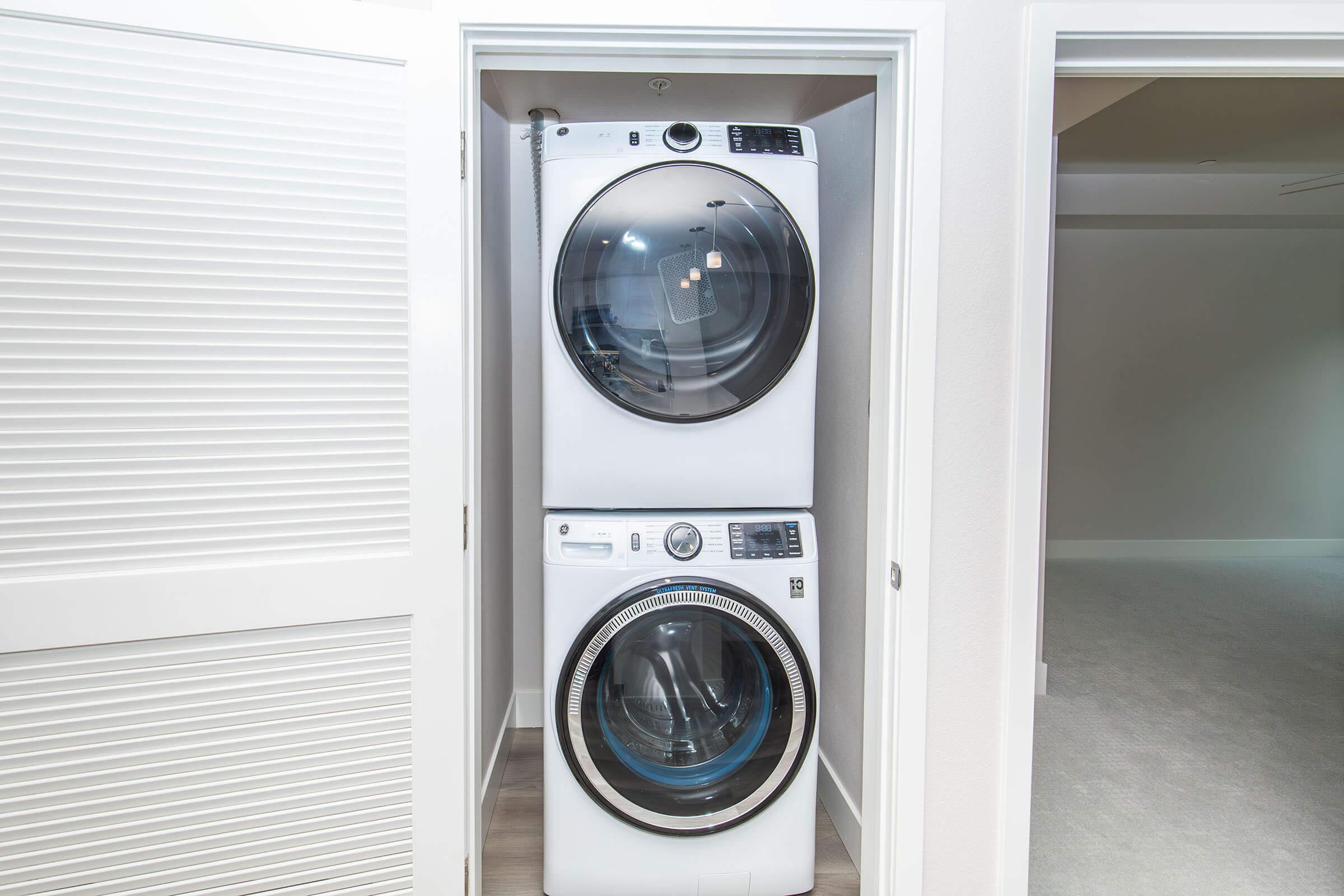 A stacked washer and dryer unit in a small laundry closet, featuring a white exterior and modern design, against a light-colored wall. The door to the closet is partially open, revealing the appliances, while a doorway is visible in the background.