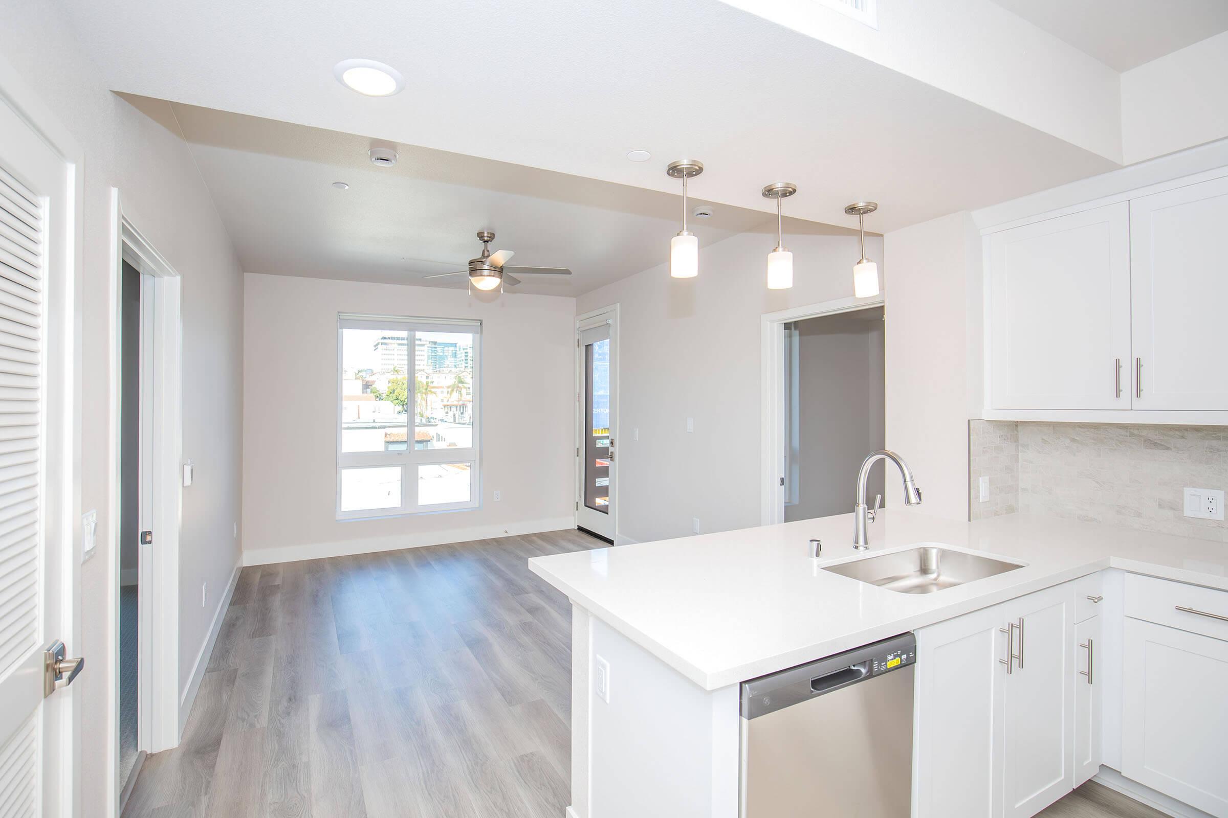 Modern kitchen and living area in an apartment, featuring a white countertop, stainless steel sink, and appliances. Large windows providing natural light, light wood flooring, and contemporary lighting fixtures enhance the open layout. A doorway leads to another room, and a ceiling fan is visible.